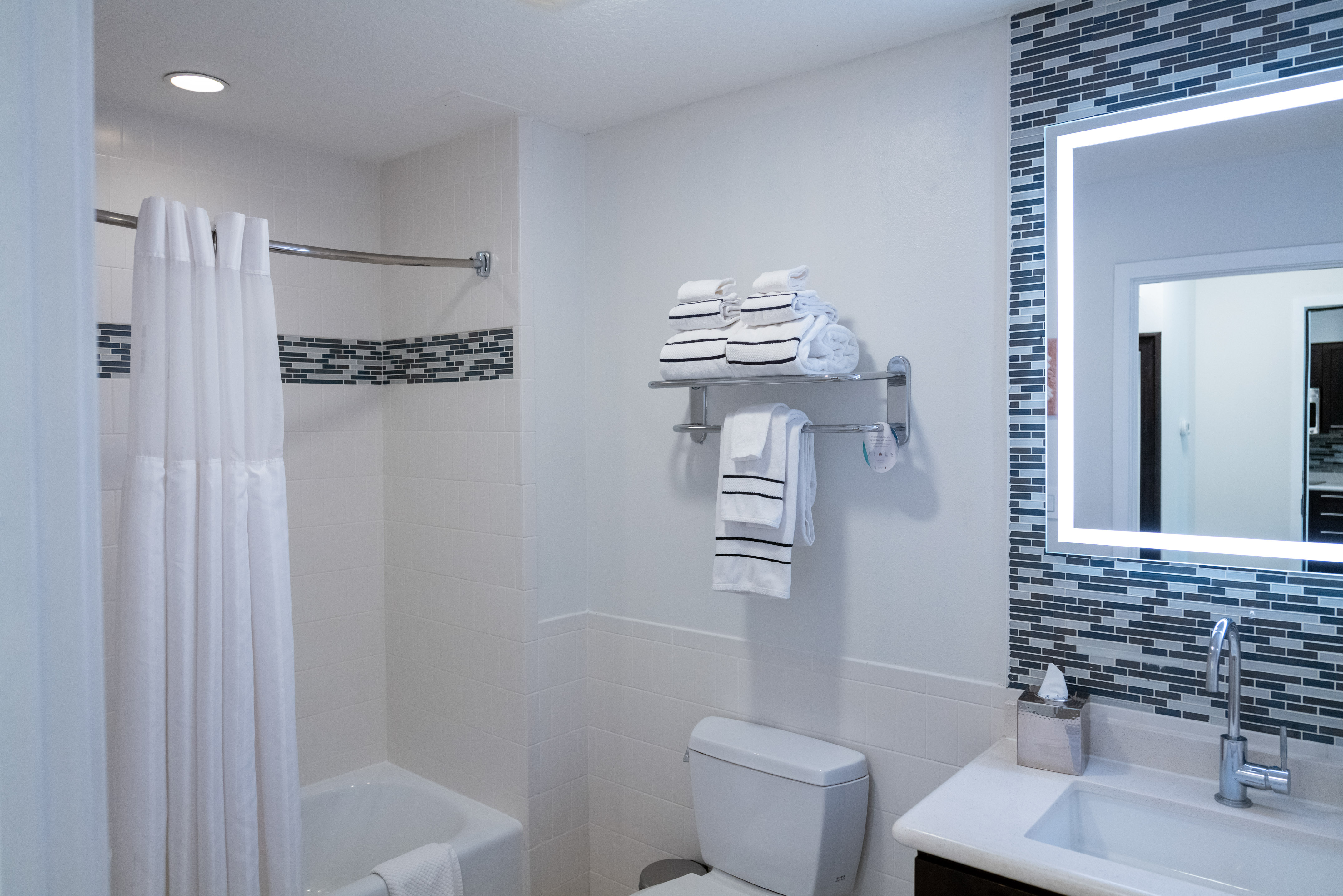 Modern bathroom with white and mosaic tiles, shower, illuminated mirror, sink.