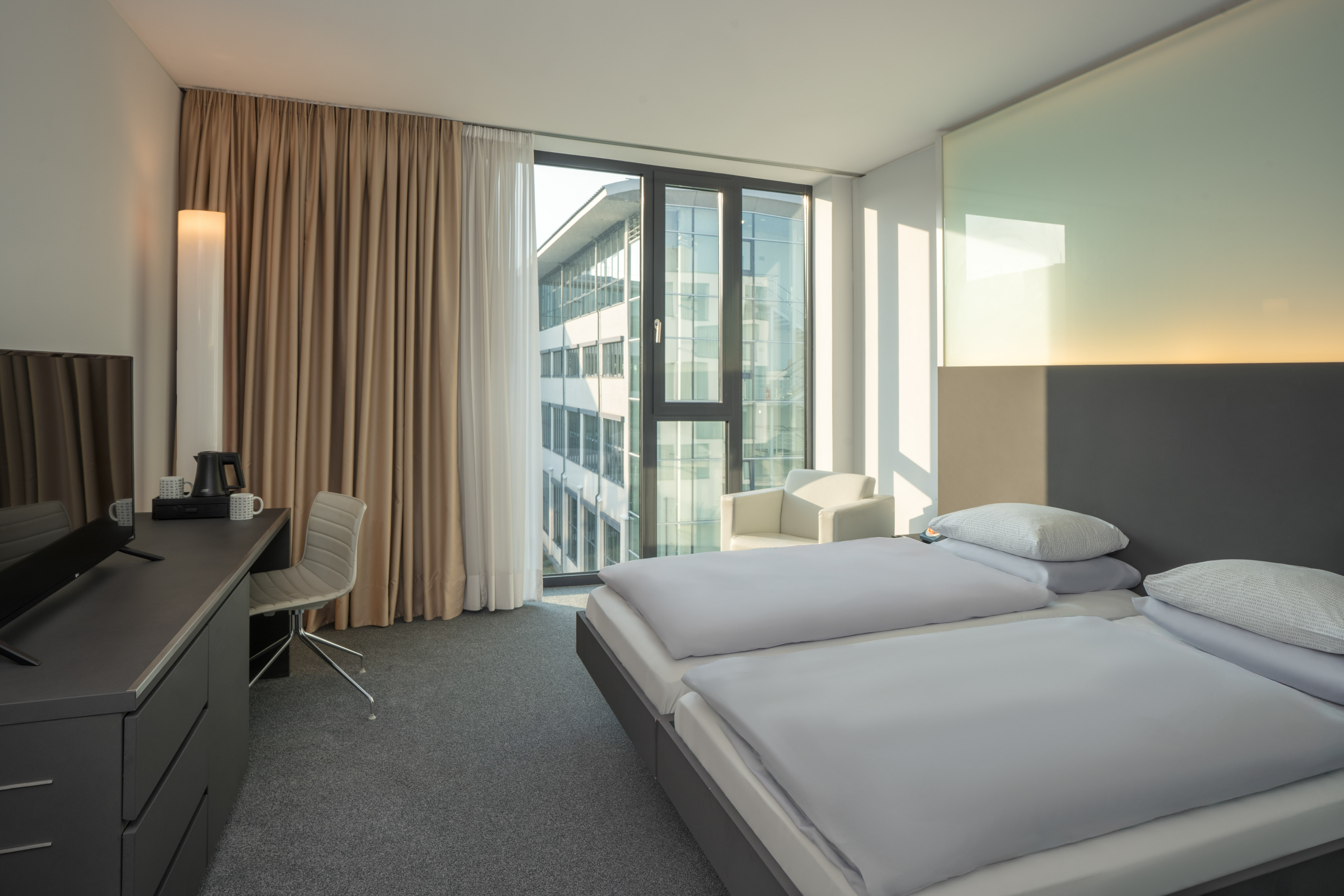 Modern hotel room with twin beds, city view, desk, and comfortable armchair.