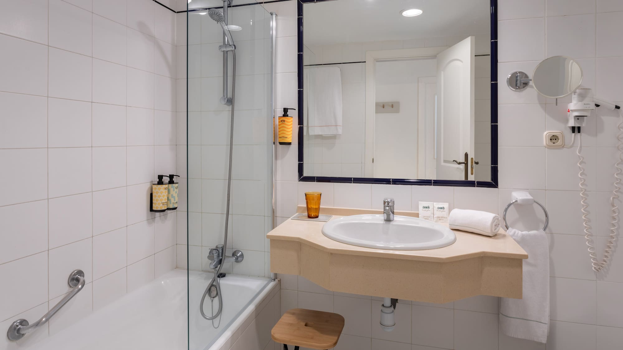 White tiled hotel bathroom with bathtub, glass shower, vanity sink, and mirror.