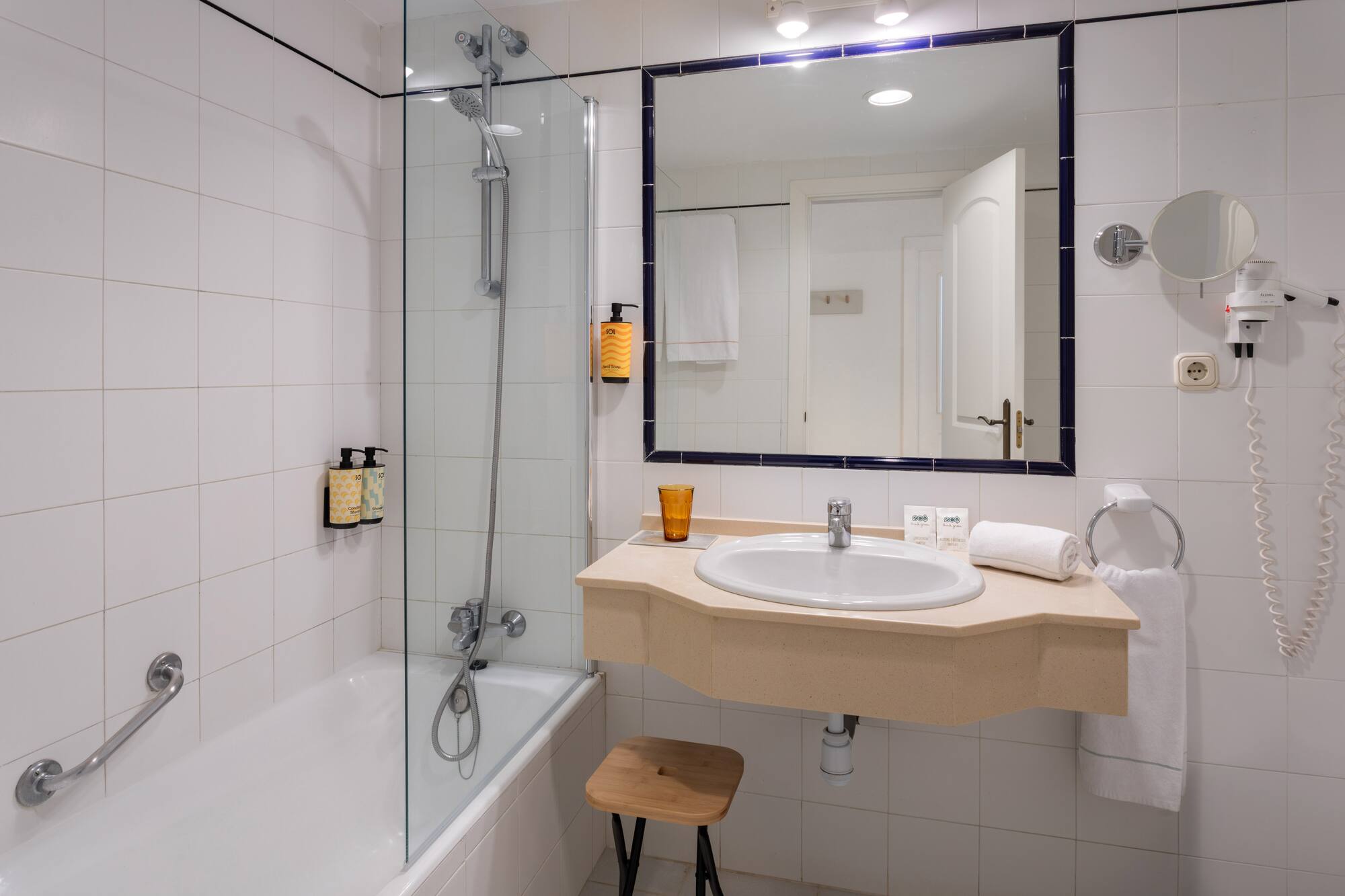 White tiled hotel bathroom with bathtub, shower, vanity sink, and mirror.