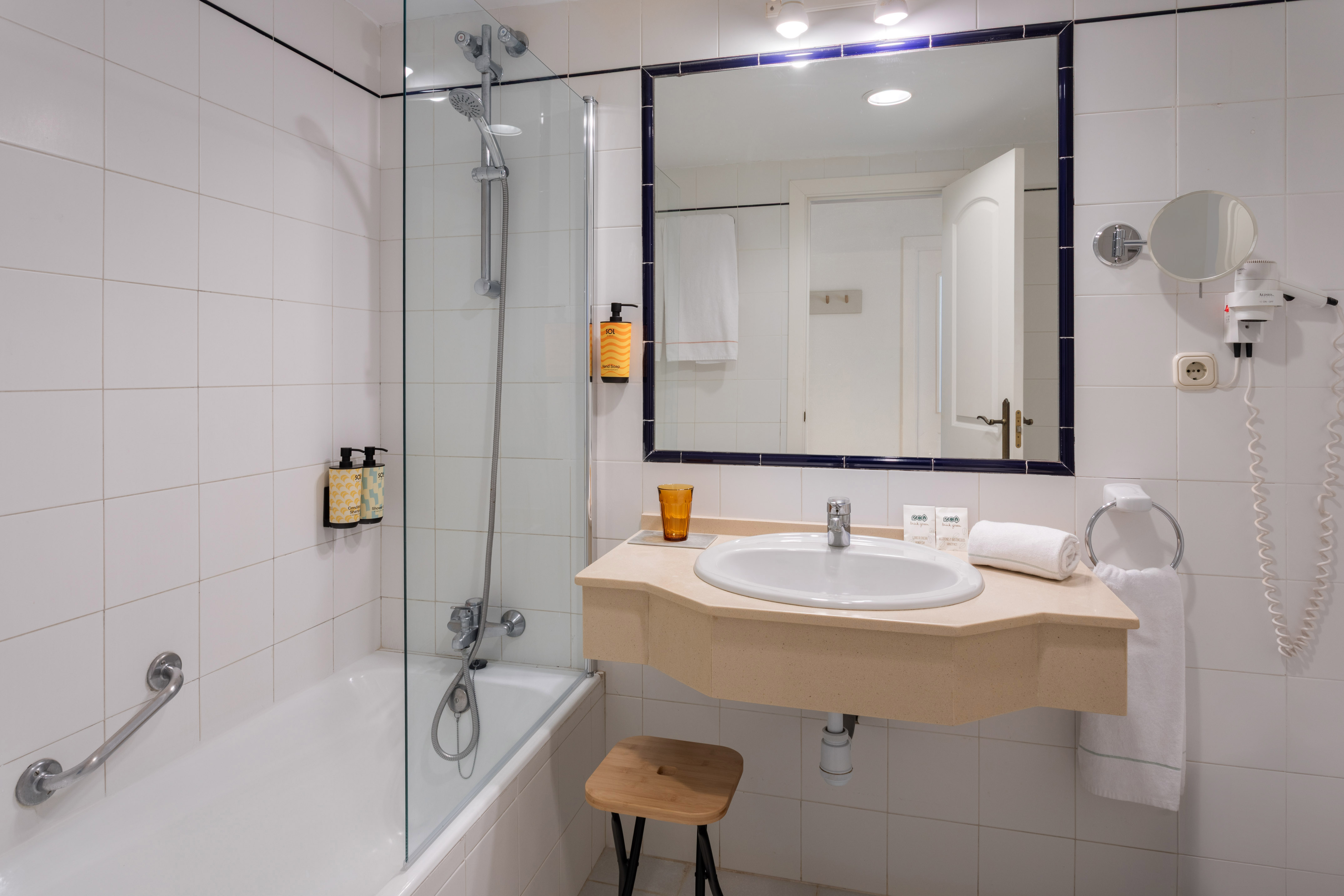 White tiled hotel bathroom with bathtub, glass shower, vanity sink, and mirror.