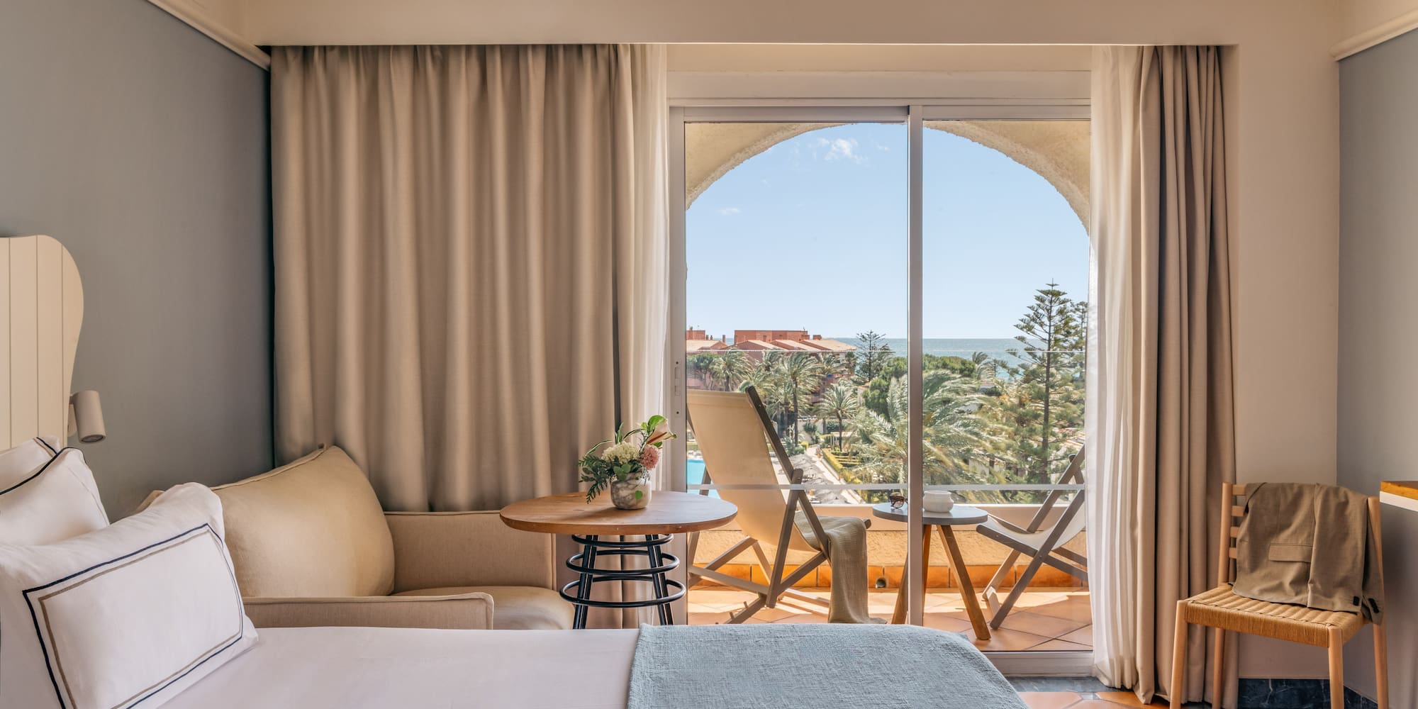 Bright hotel room with bed, balcony, and expansive sea view over a resort.