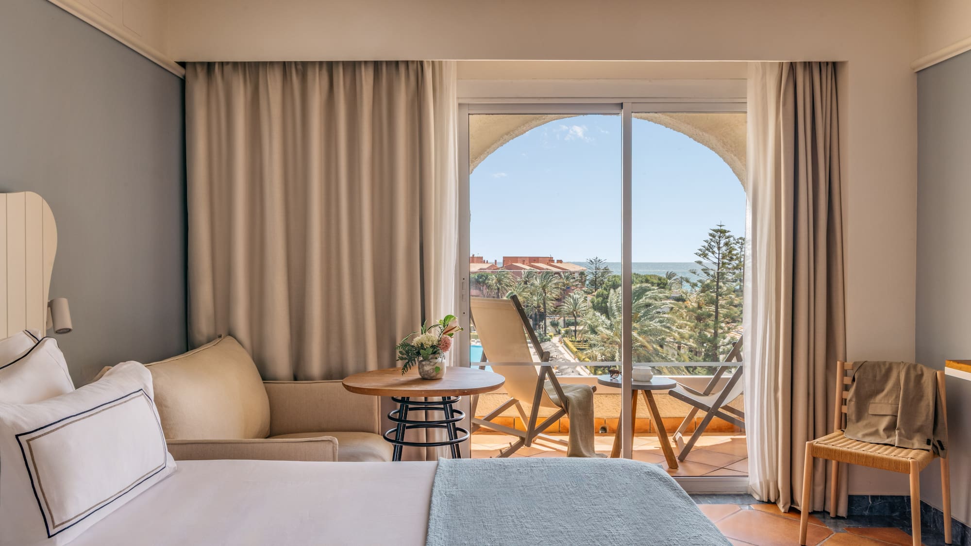 Bright hotel room with bed, balcony, and expansive sea view over a resort.