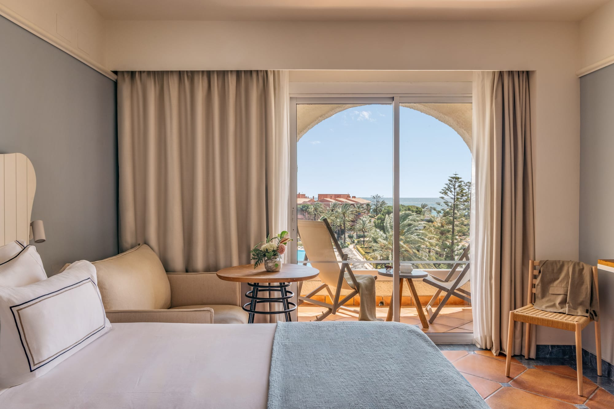 Bright hotel room with bed, balcony, and expansive sea view over a resort.