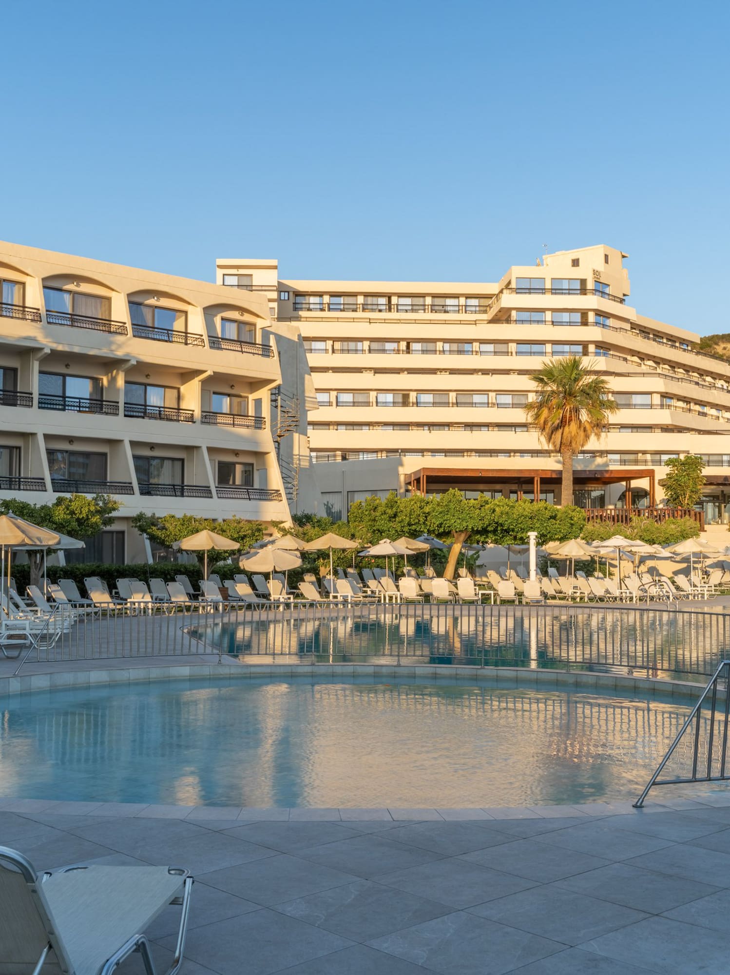 Large hotel resort with a round swimming pool, sun loungers, and palm trees.