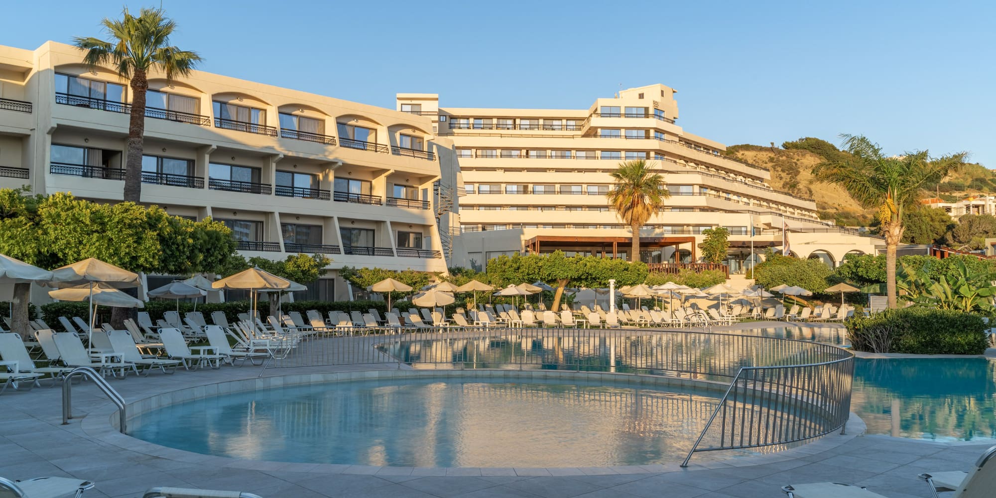 Sunny hotel resort with large outdoor pool, lounge chairs, and palm trees.
