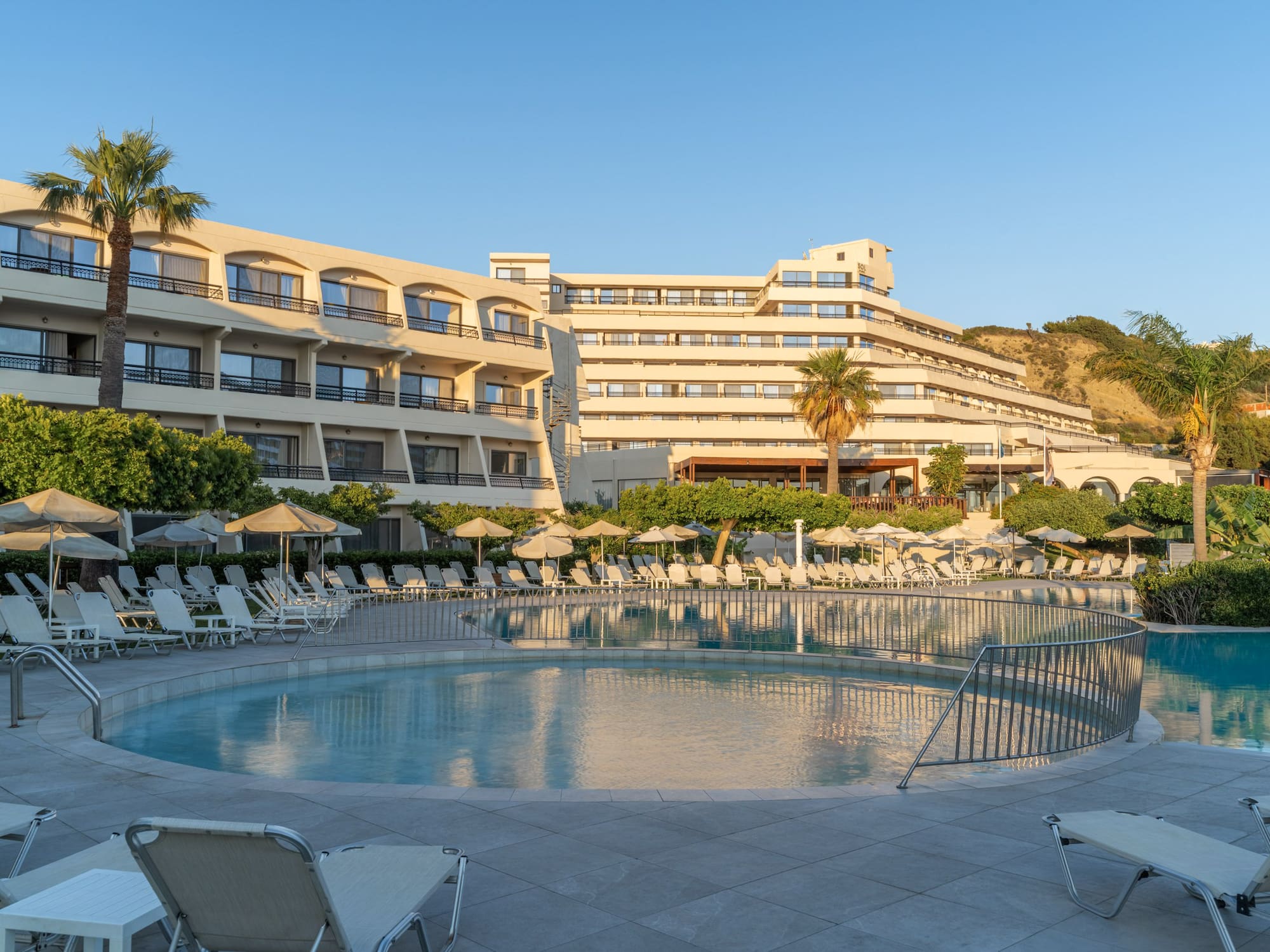 Sunny resort pool with hotel, sun loungers, and tropical landscaping.