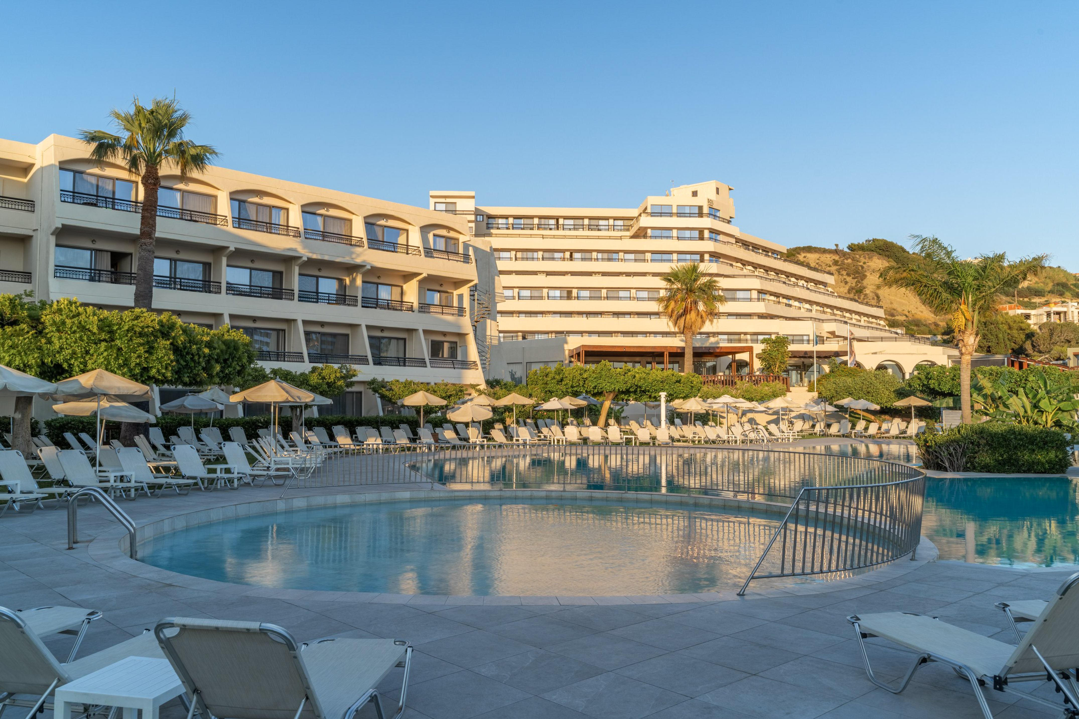 Large hotel resort with a round swimming pool, sun loungers, and palm trees.