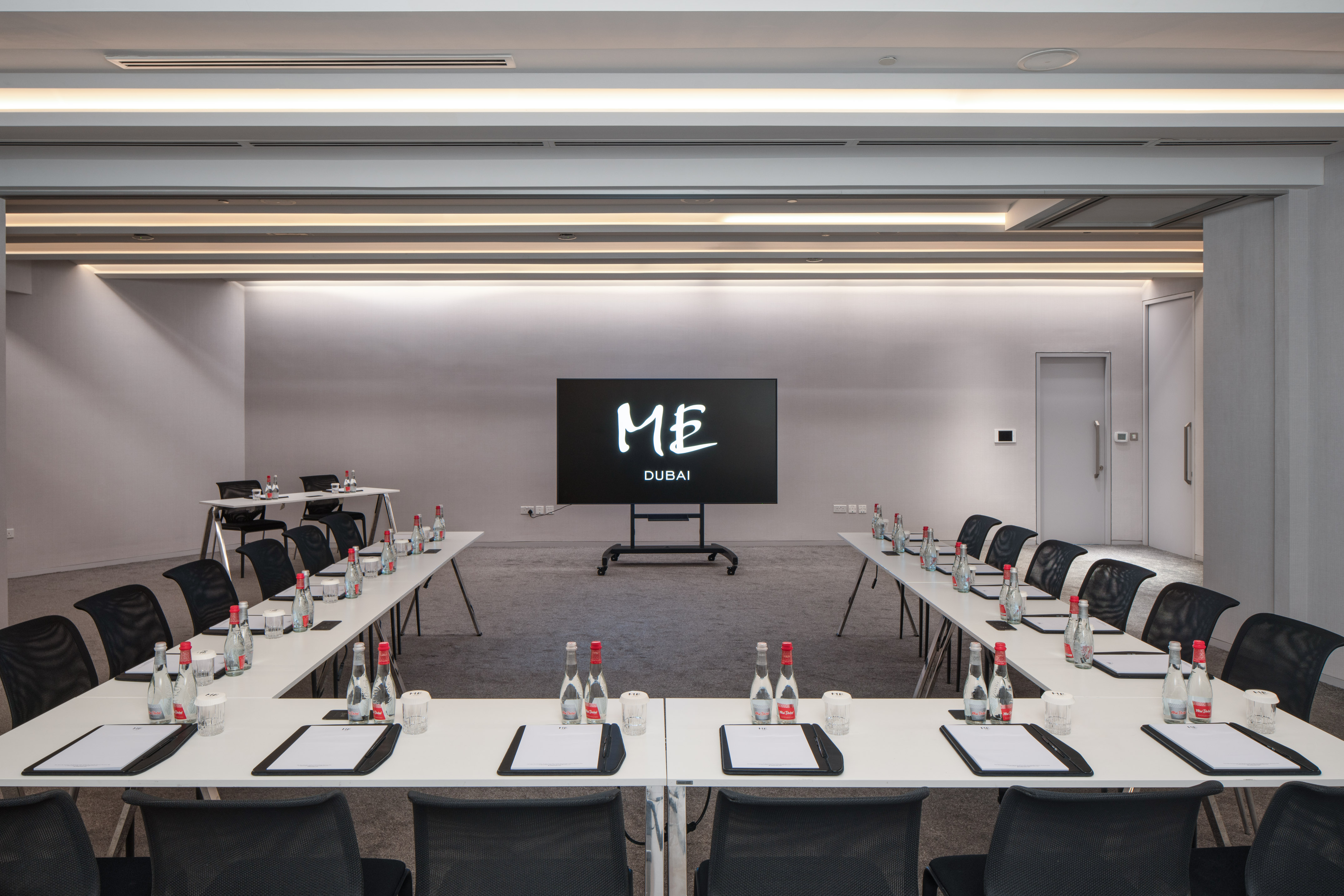 Modern ME Dubai meeting room with U-shaped tables, screen, and water.