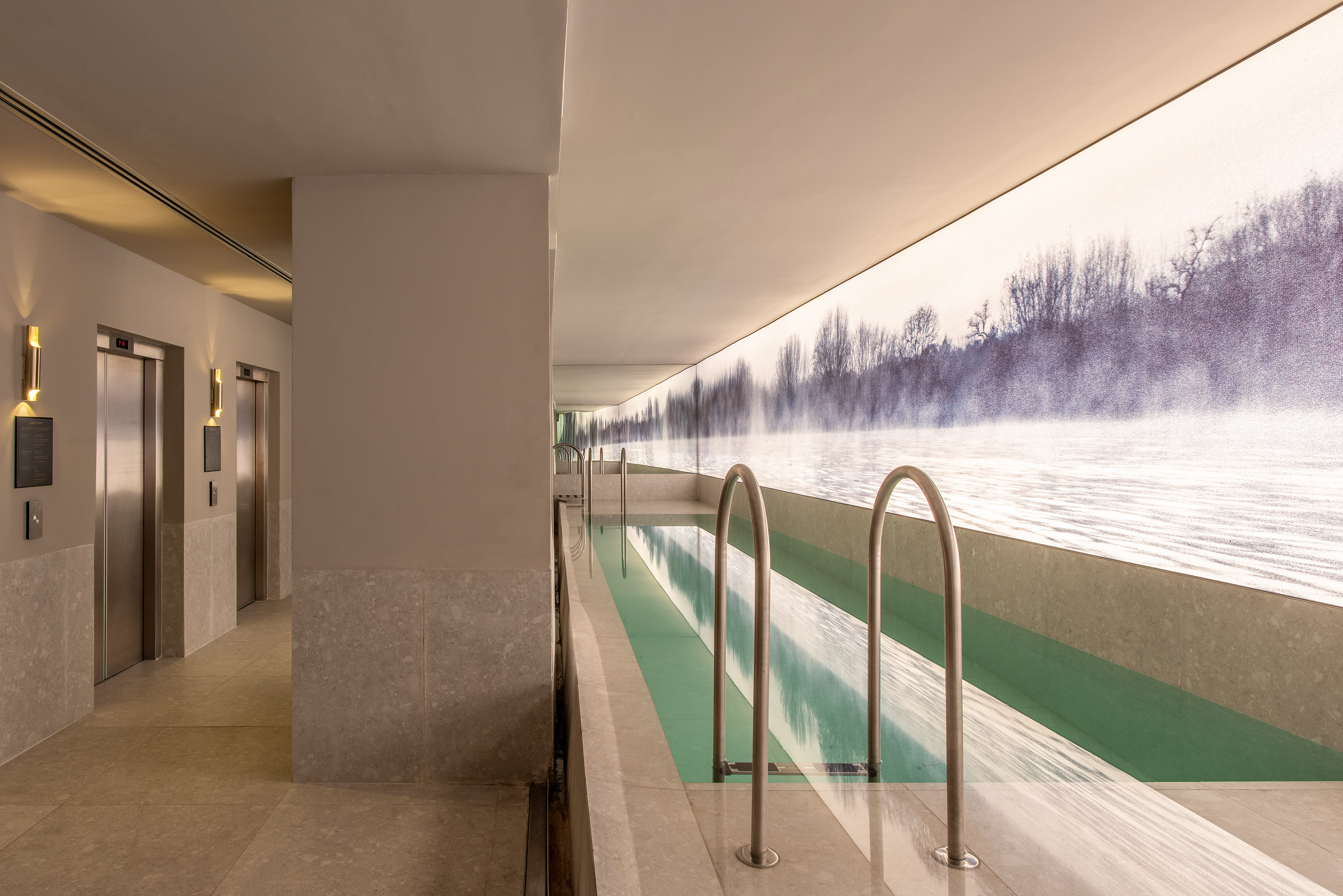 Sleek indoor lap pool with illuminated misty forest mural and elevators.
