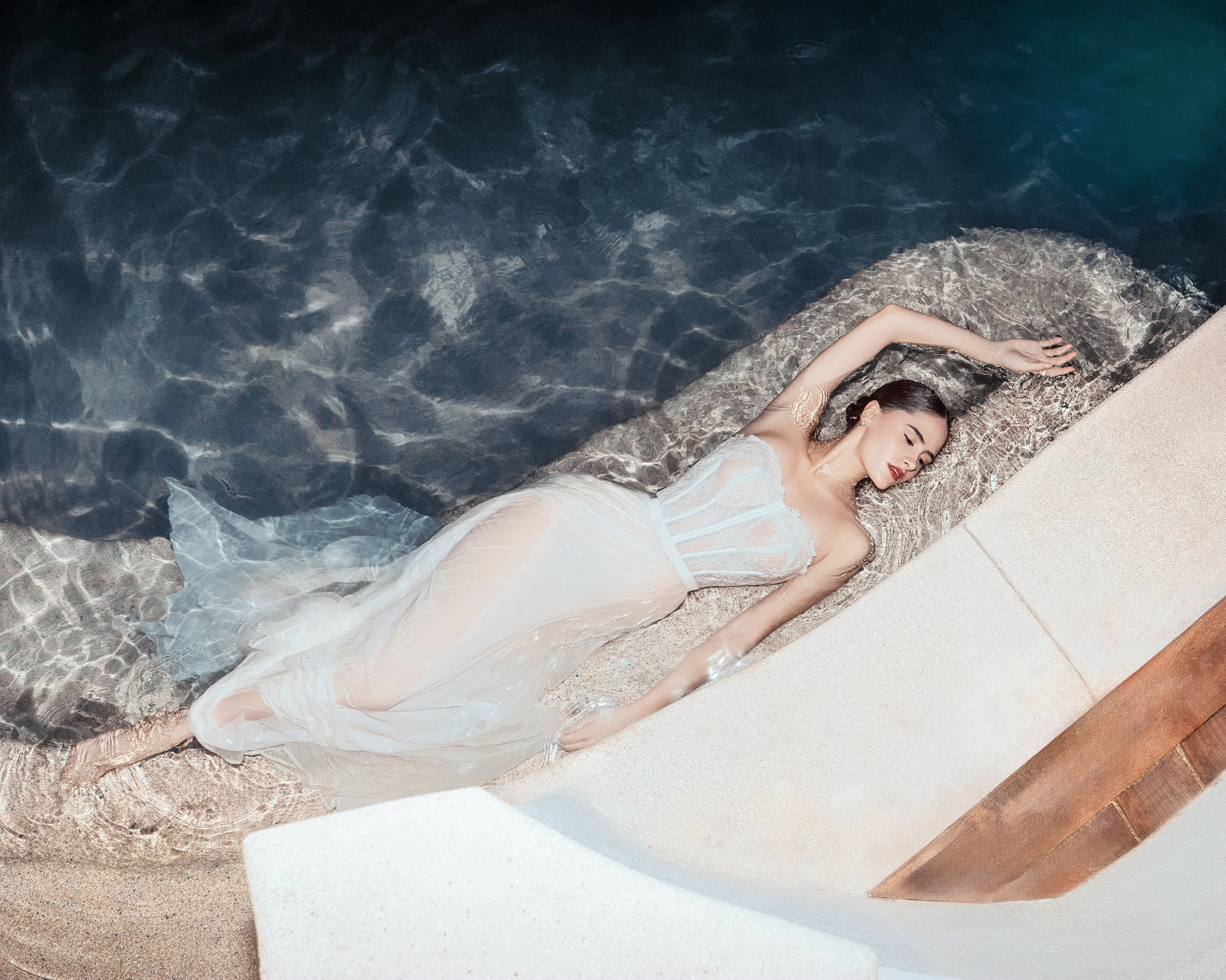 Woman in white lace dress lying serenely in clear pool water.