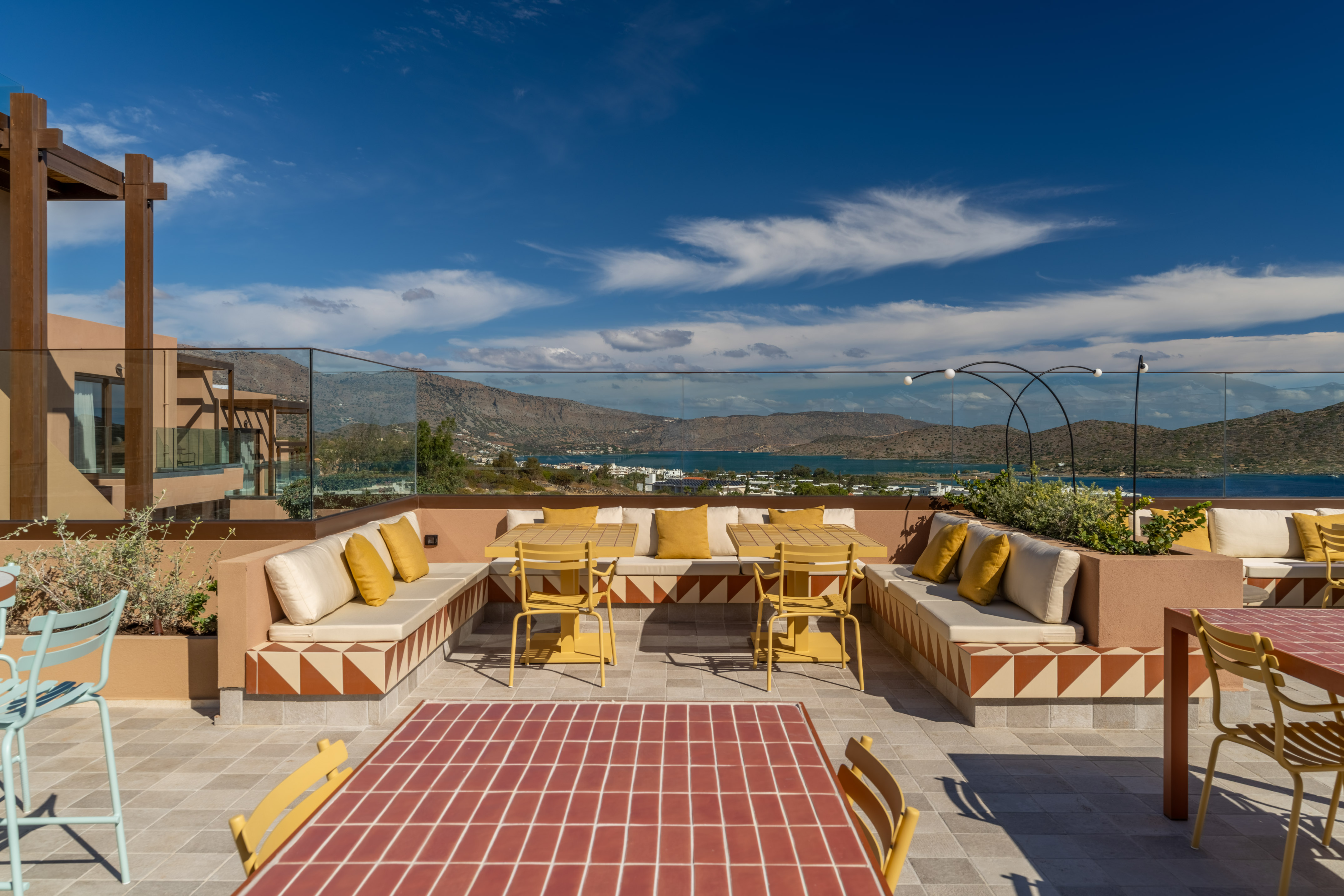 Rooftop terrace with yellow outdoor seating, geometric patterns, sea, and mountain views.