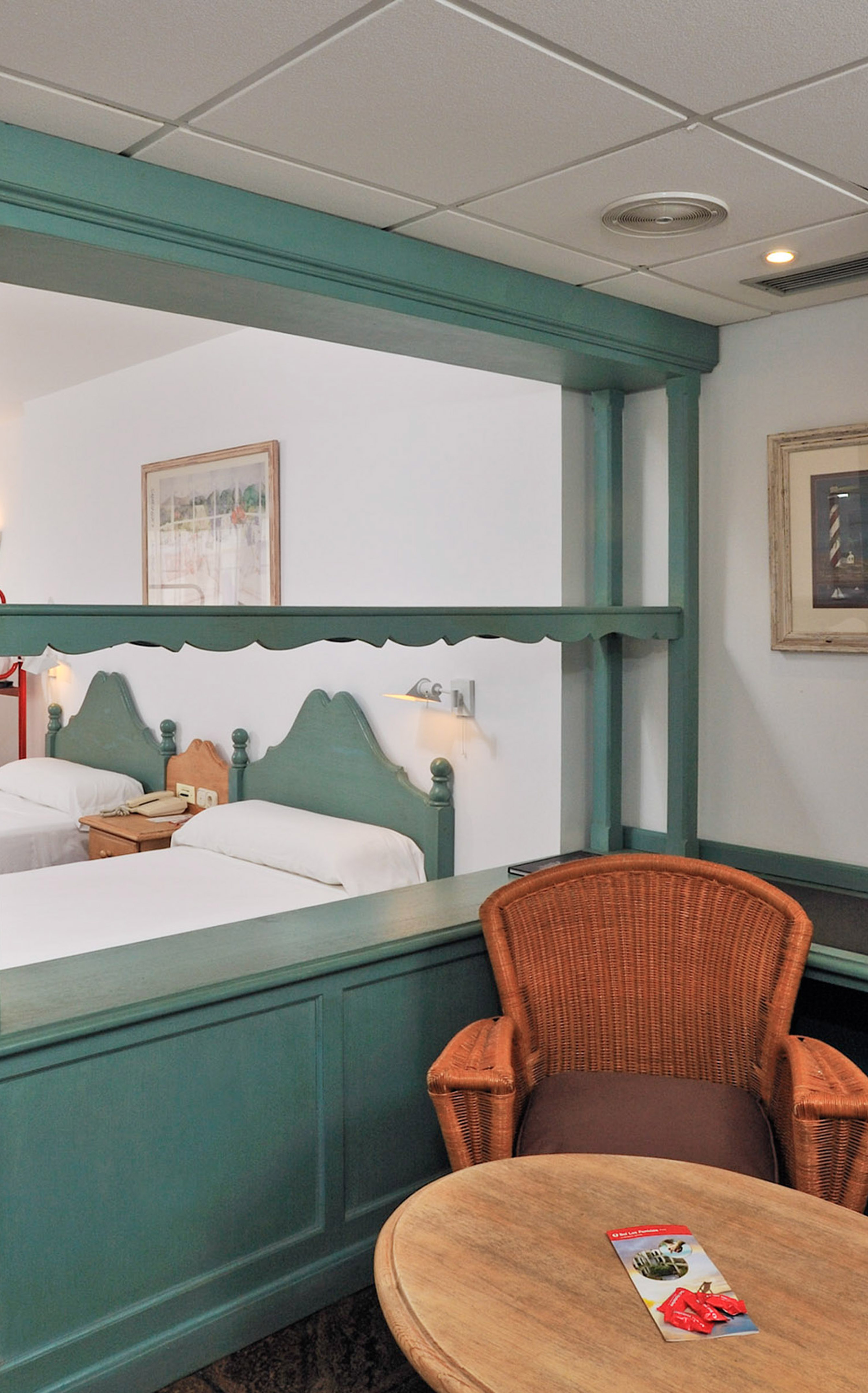 Hotel room with twin beds, green partition, wicker chair, and wooden side table.