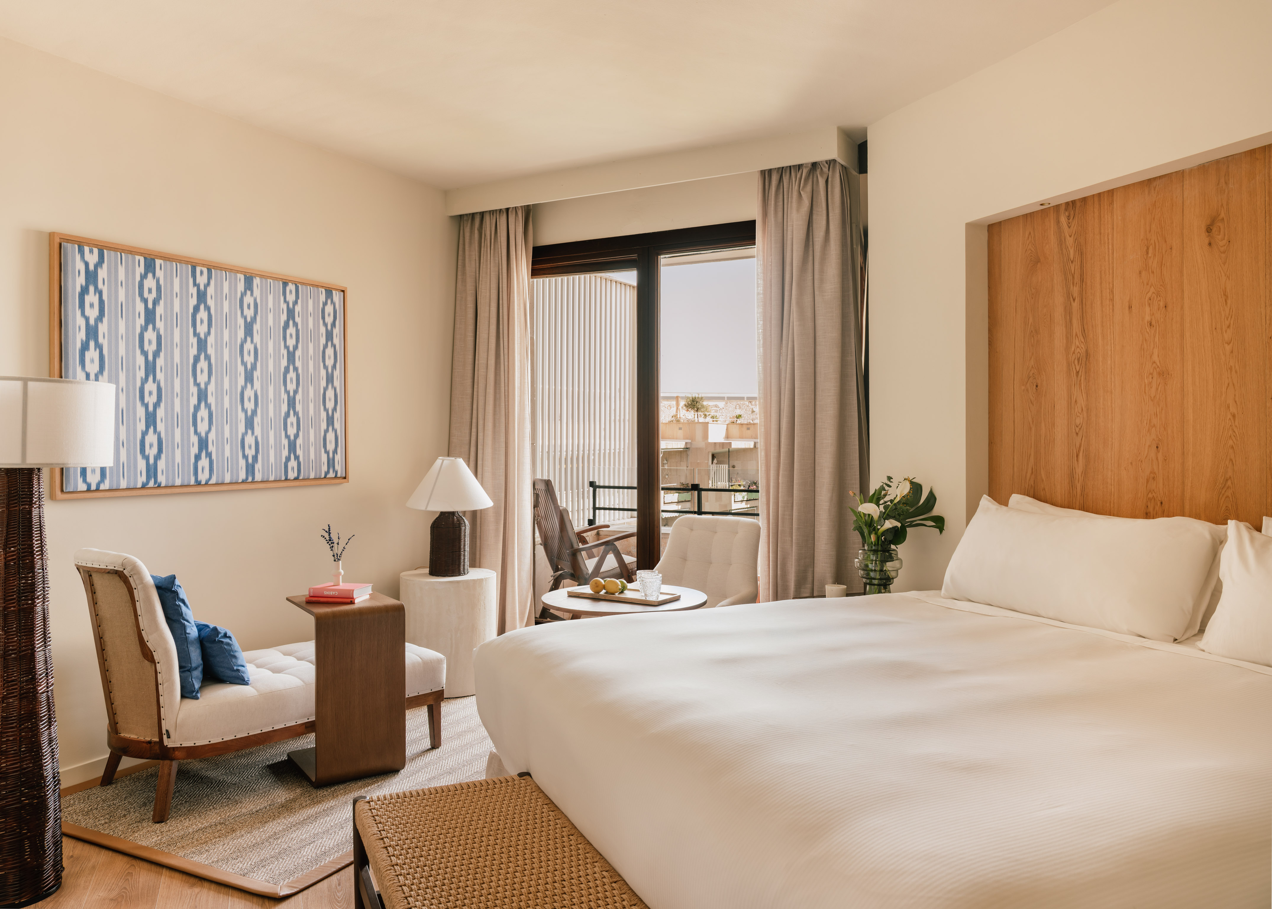 Hotel room with king bed, balcony, wooden headboard, lounge chair, and blue art.
