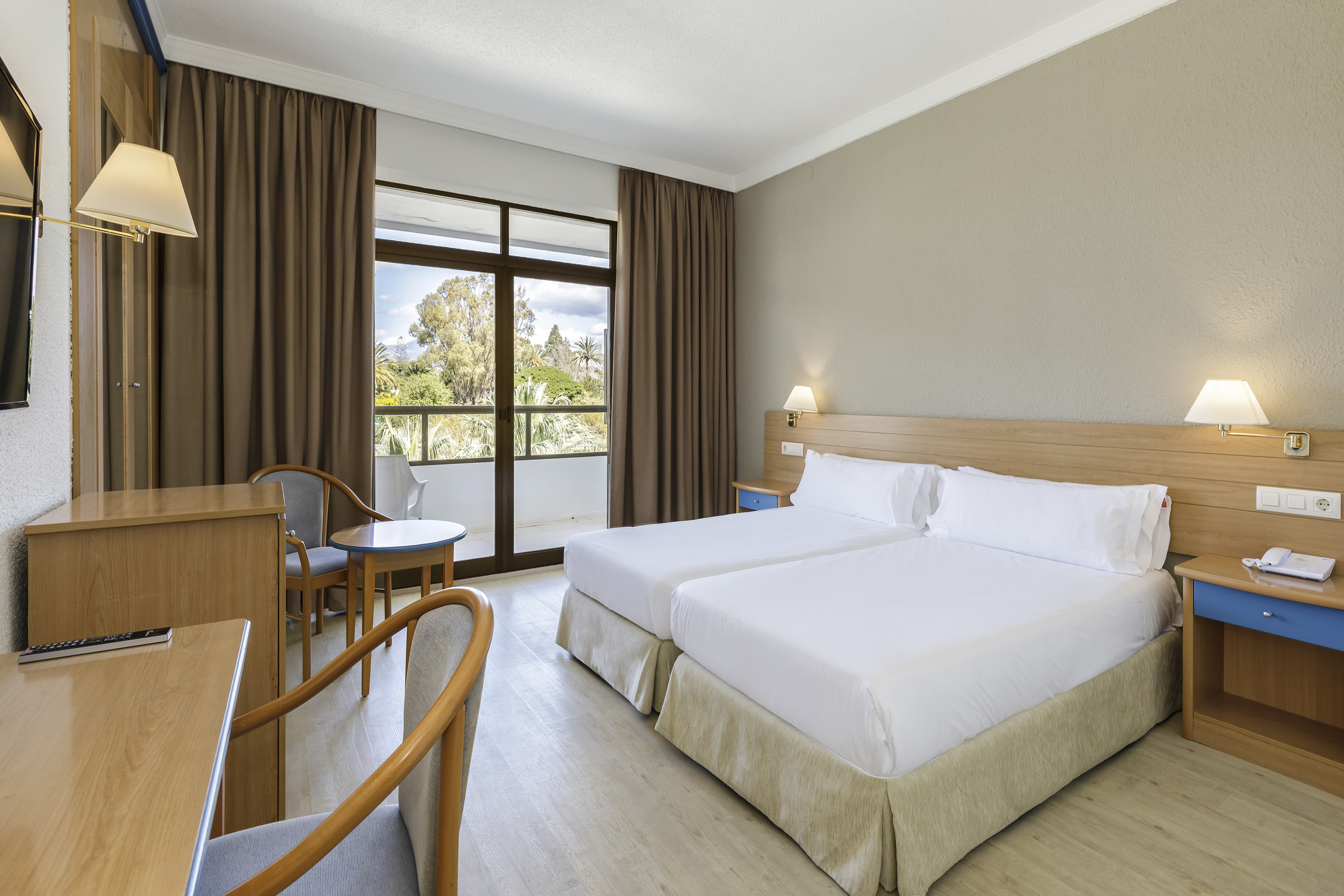 Hotel room with twin beds, wooden furniture, balcony, and garden view.