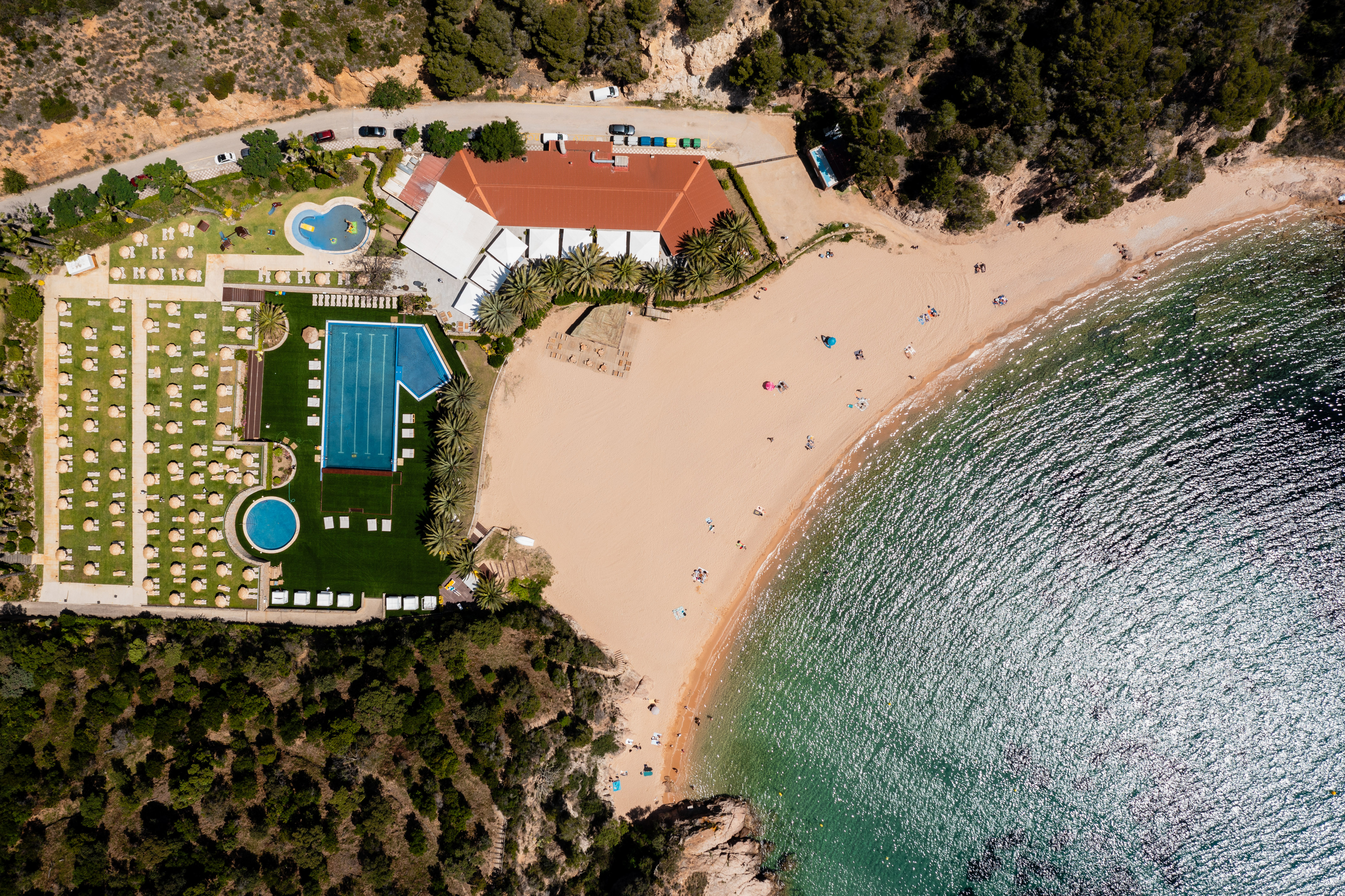 Aerial view: Sandy beach resort with pools, palm trees, and clear blue sea.