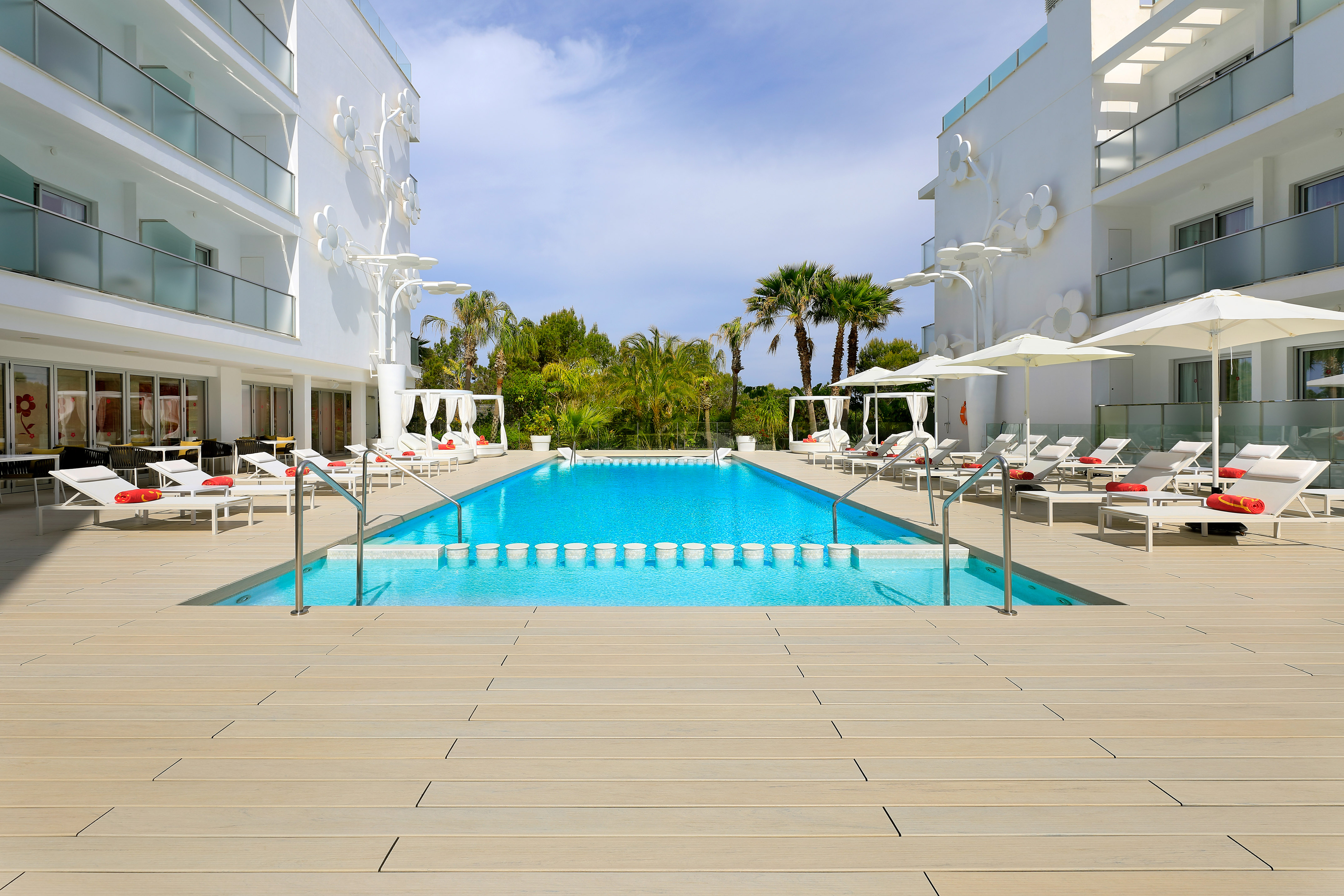 Modern resort pool, white buildings, wooden deck, sun loungers, palm trees.