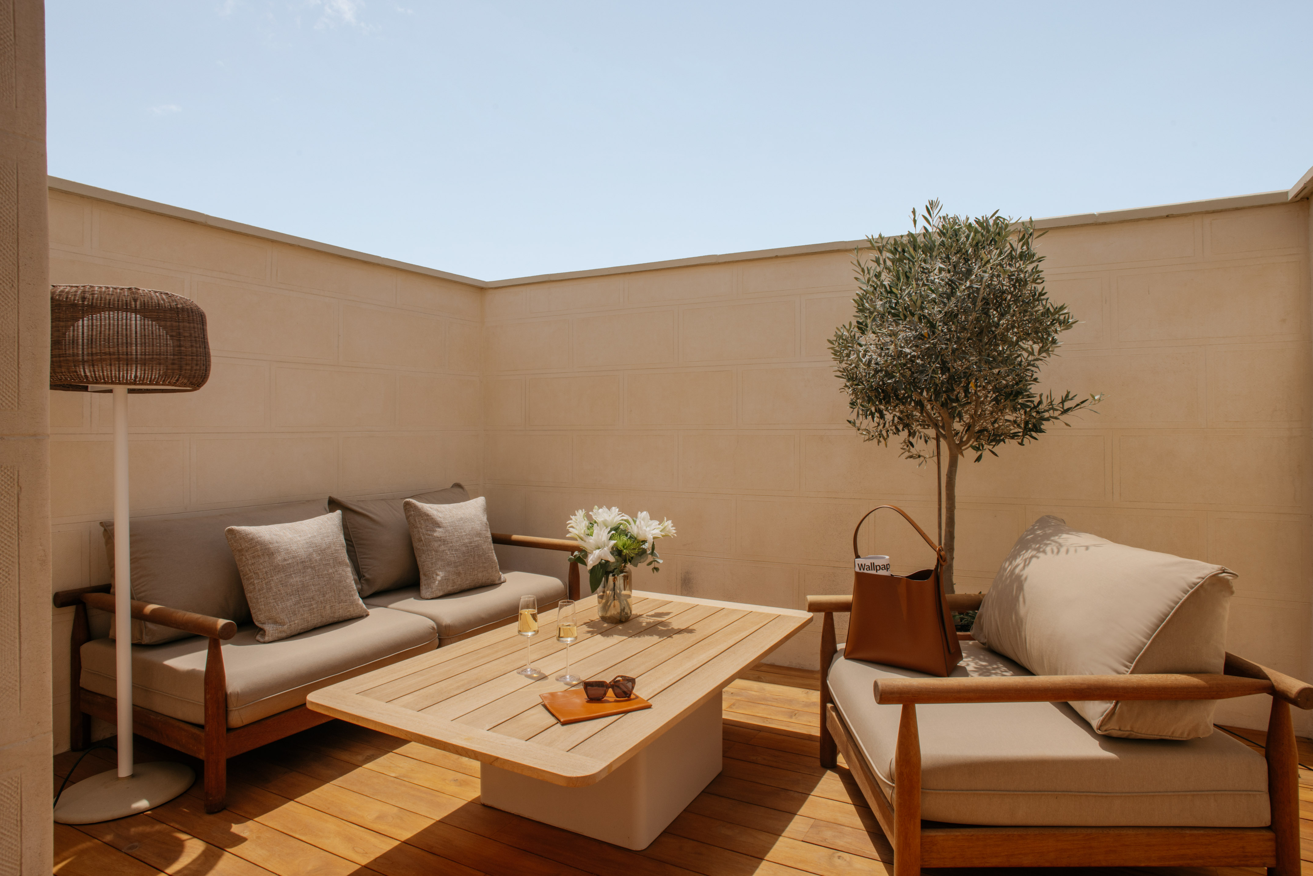 Modern outdoor patio lounge with wooden furniture, beige cushions, and an olive tree.