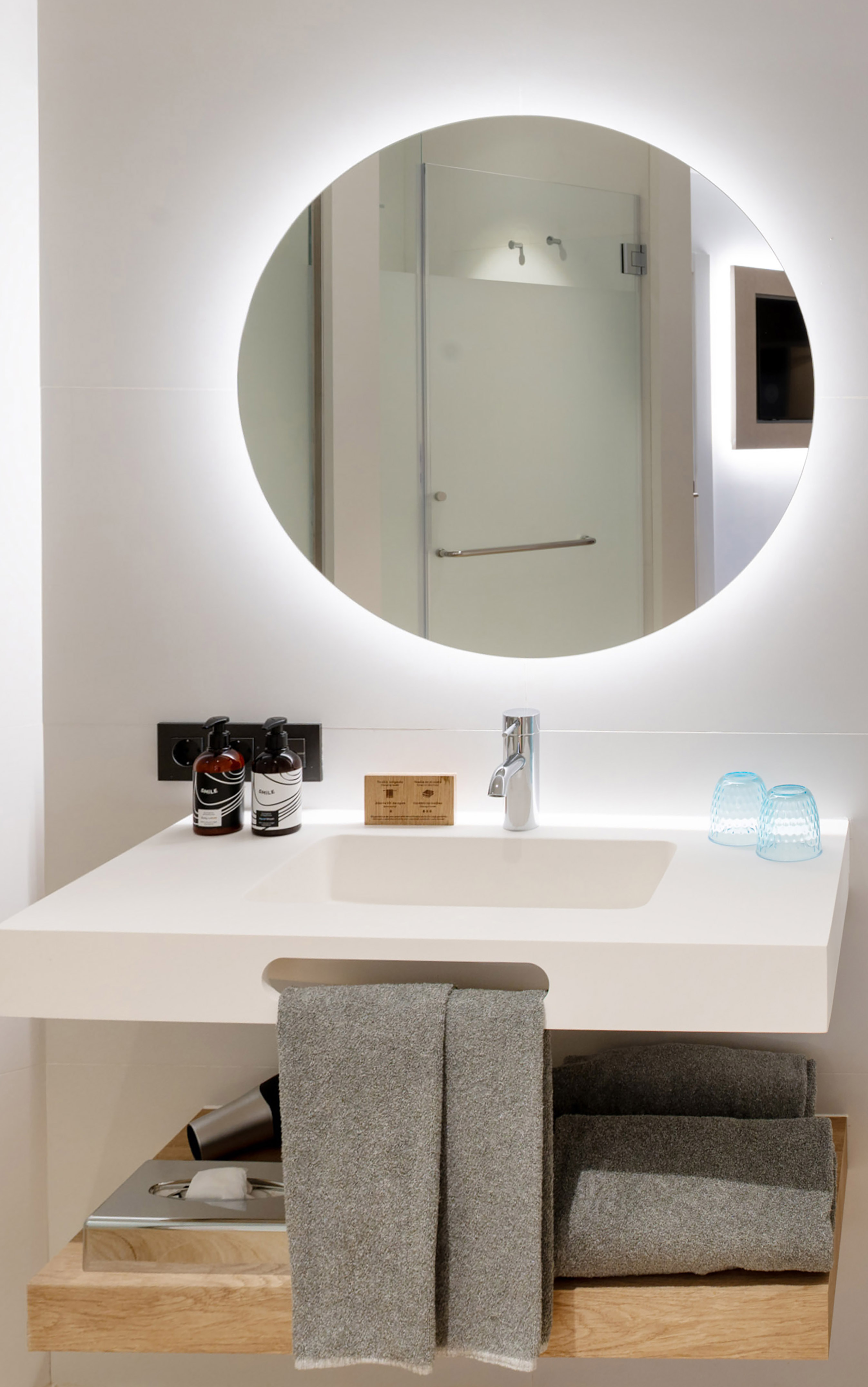 Modern bathroom vanity with backlit mirror, sink, toiletries, and gray towels.