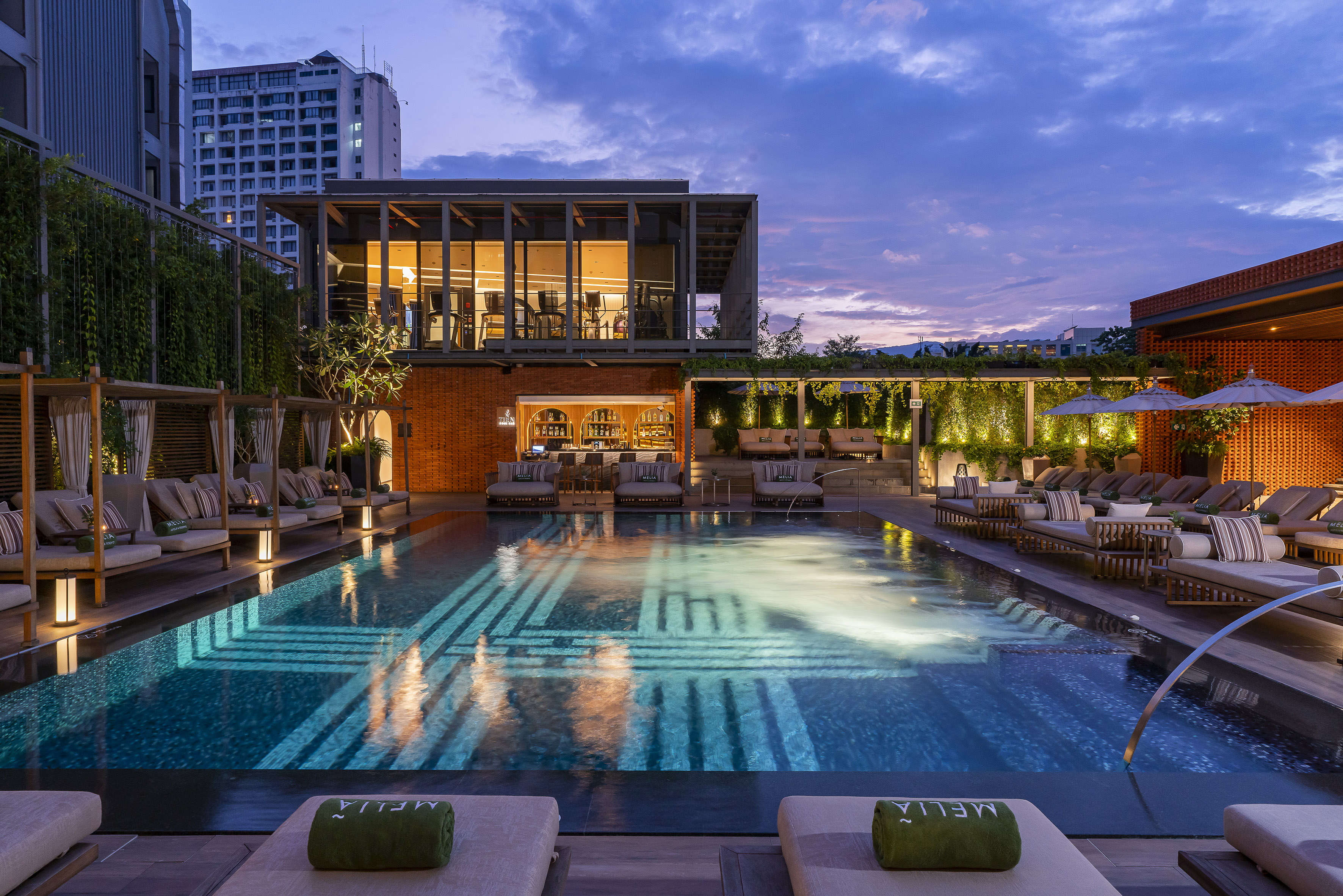 Meliá hotel pool and bar at dusk, with glowing water, lounge chairs, and city.