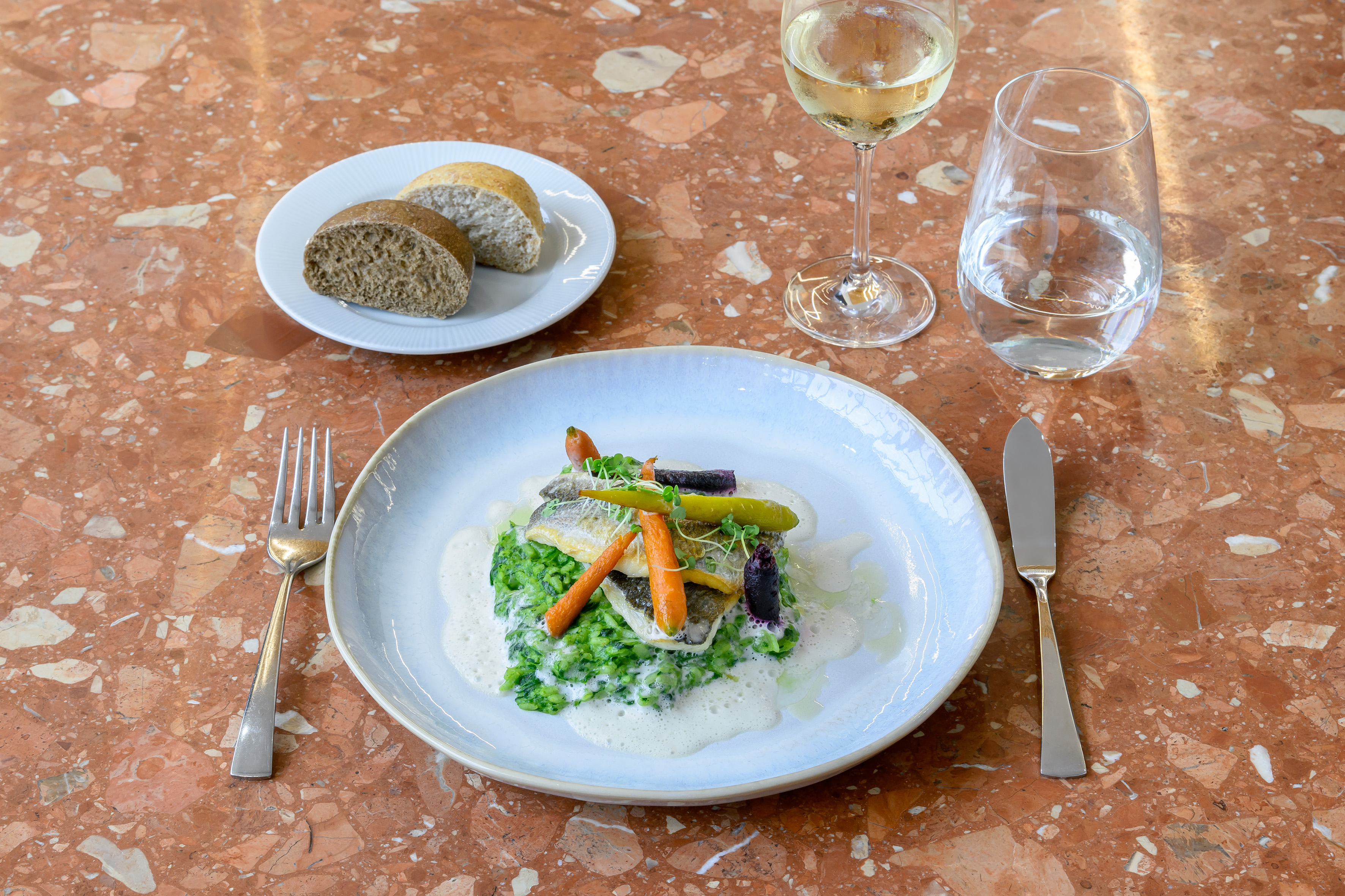 Gourmet fish and vegetable entree with wine and bread on a marble table.