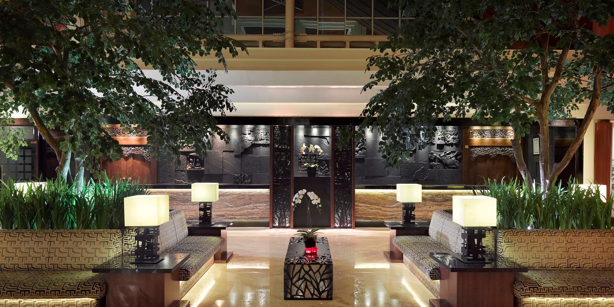 Elegant hotel lounge with lush trees, symmetrical seating, and ornate carved walls.