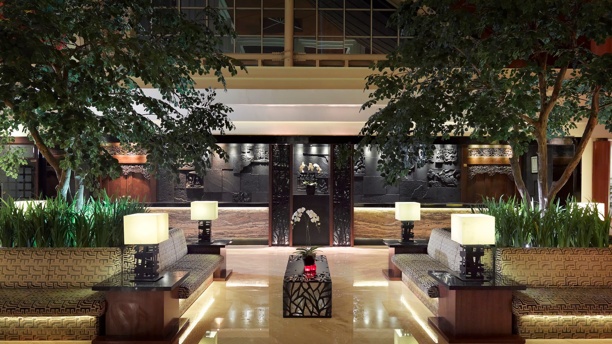 Elegant hotel lounge with lush trees, symmetrical seating, and ornate carved walls.