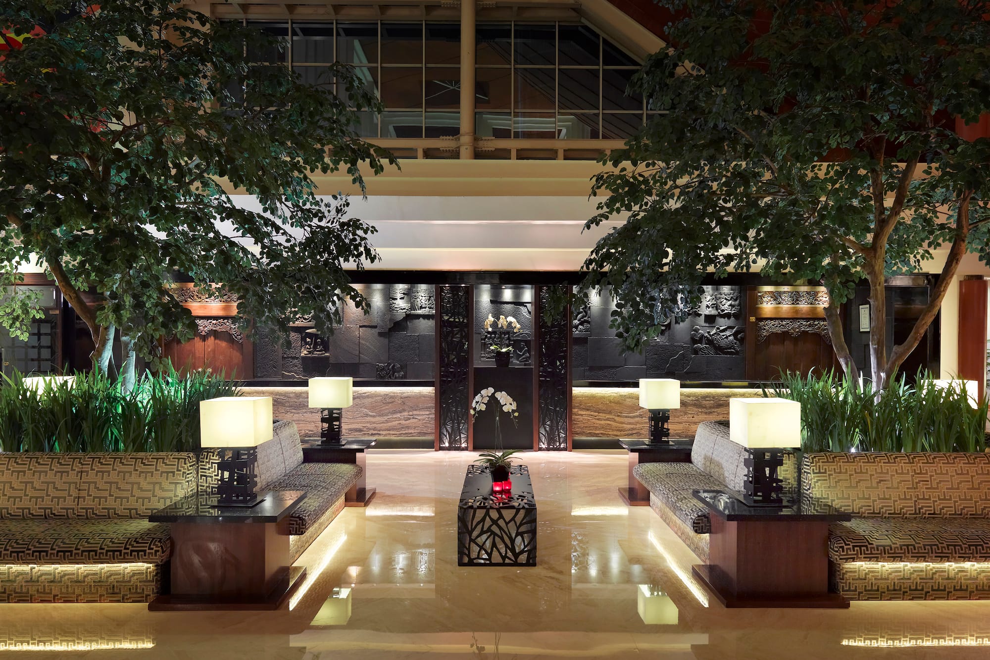 Elegant hotel lounge with lush trees, symmetrical seating, and ornate carved walls.