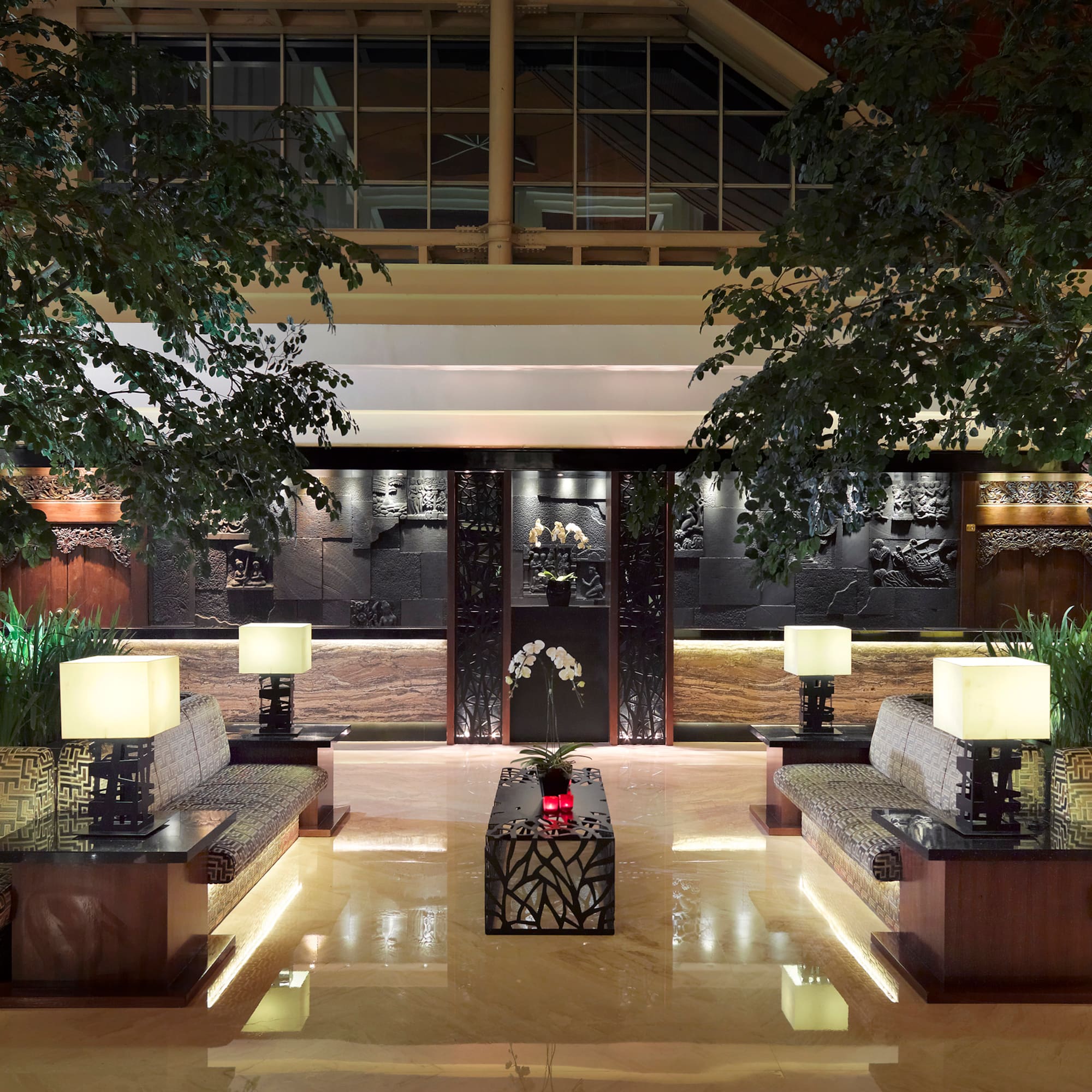 Elegant hotel lounge with lush trees, symmetrical seating, and ornate carved walls.