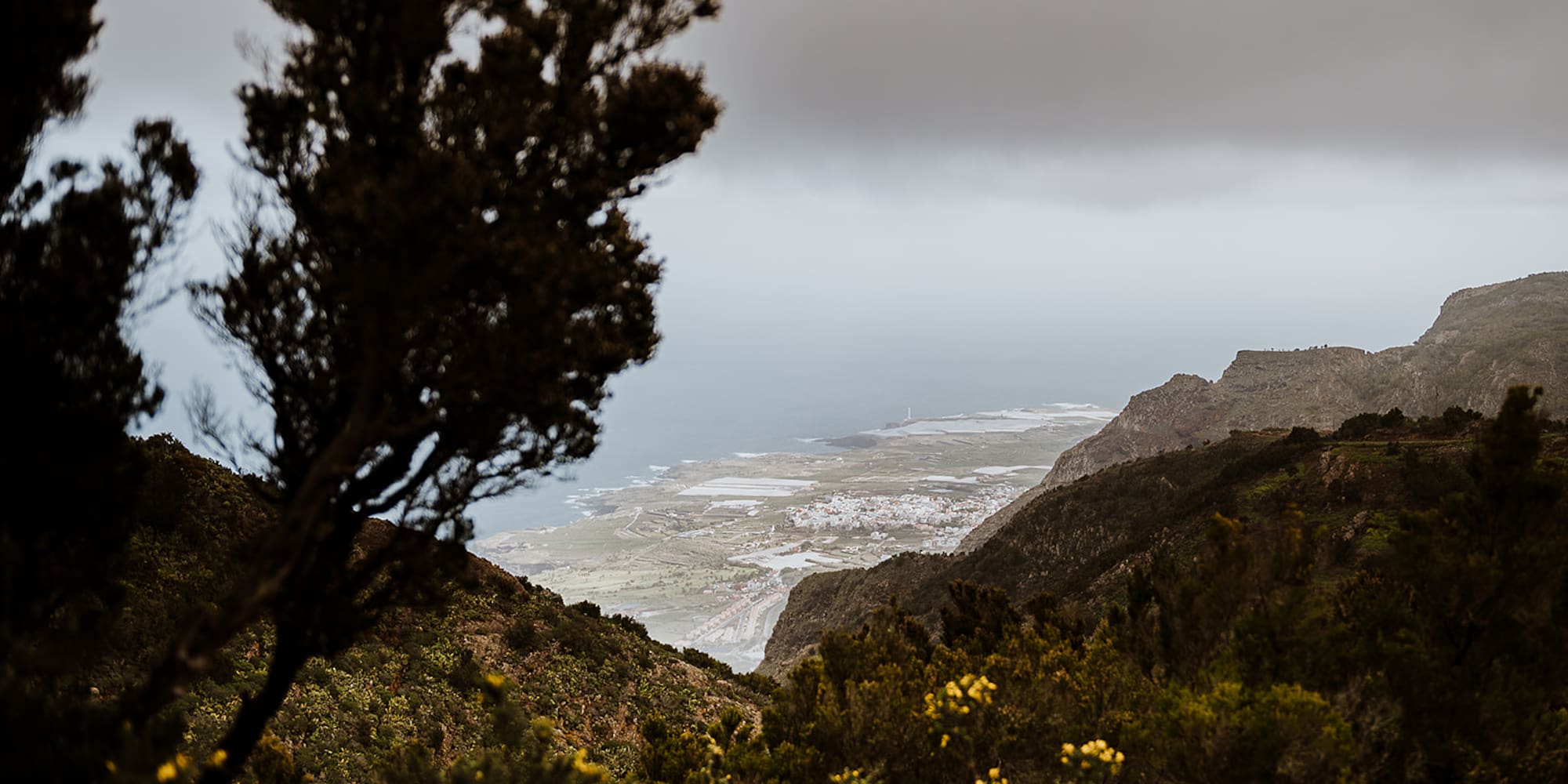 Mountain vista of coastal village, ocean, lighthouse, and green slopes under grey sky.