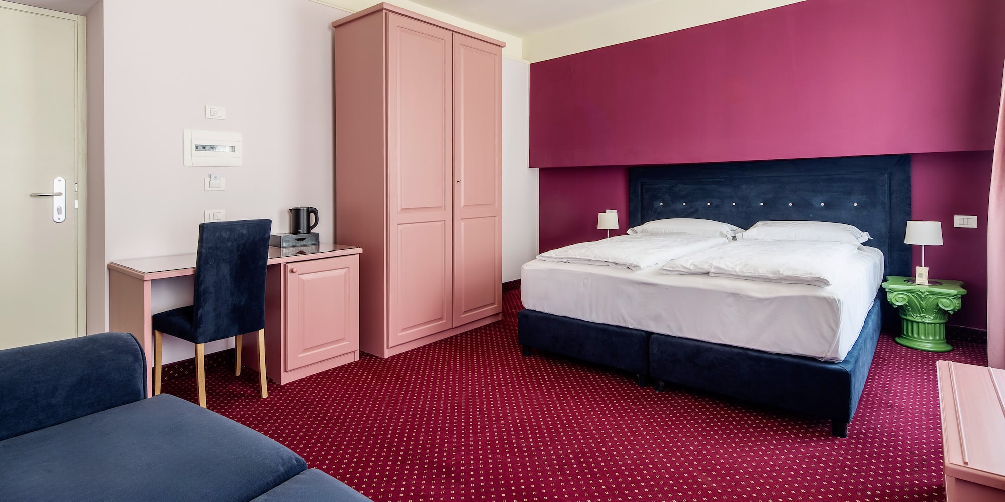 Magenta and pink hotel room with blue bed, red carpet, desk, and sofa.