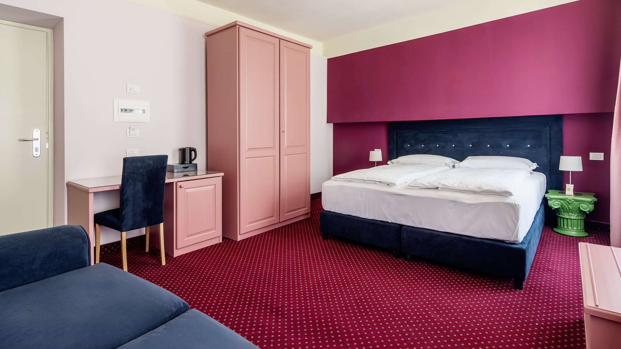 Hotel bedroom with magenta wall, blue bed, pink wardrobe, red carpet, and blue couch.