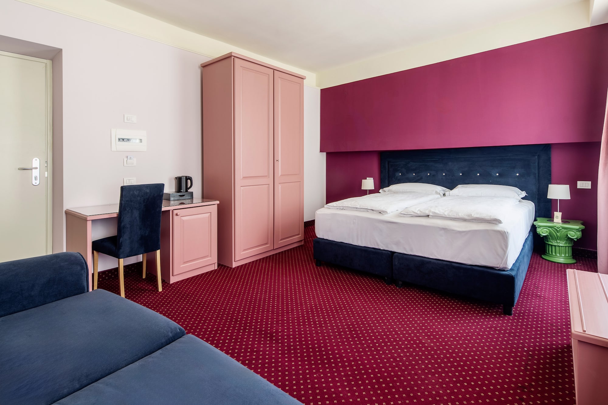 Magenta and pink hotel room with blue bed, red carpet, desk, and sofa.