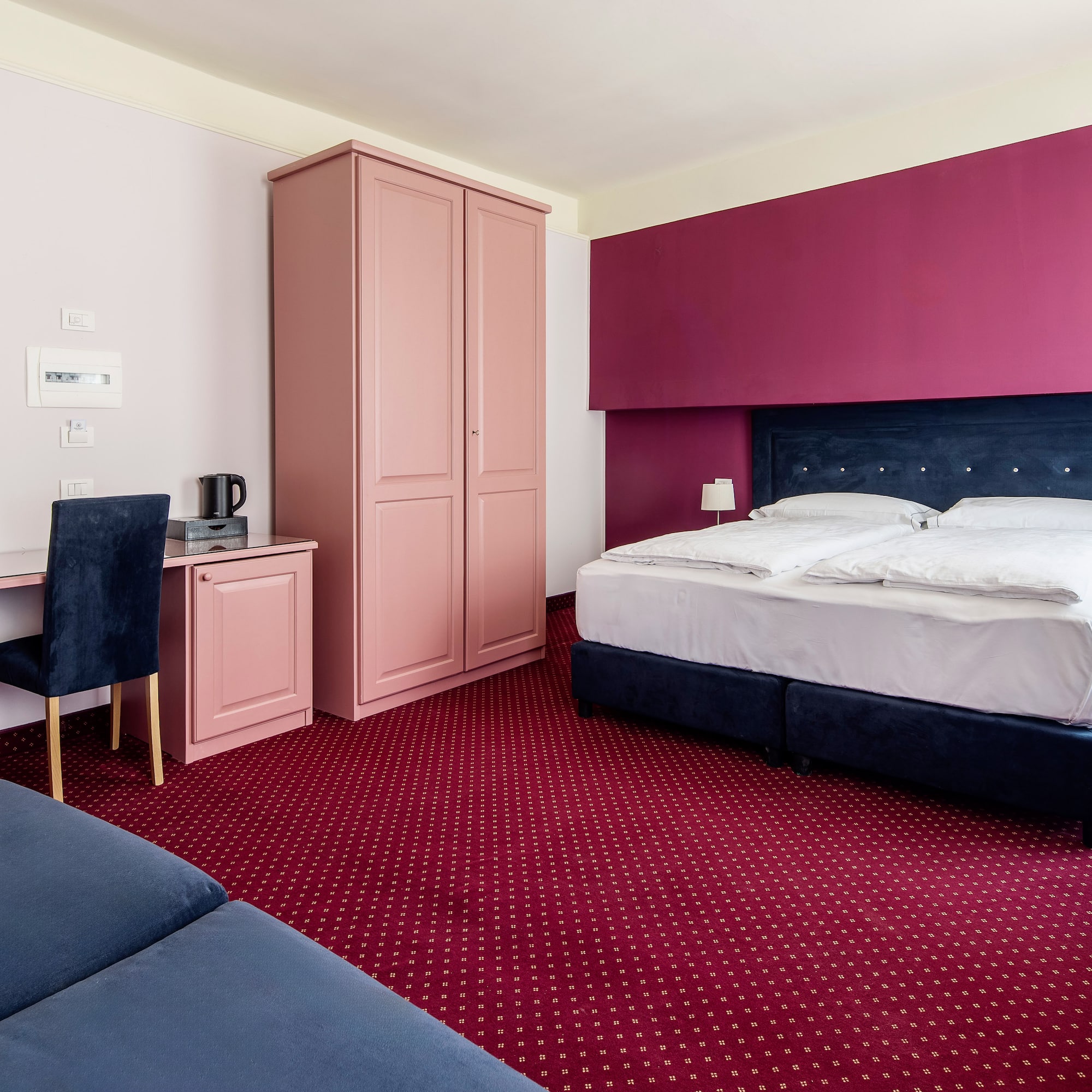 Magenta and pink hotel room with blue bed, red carpet, desk, and sofa.