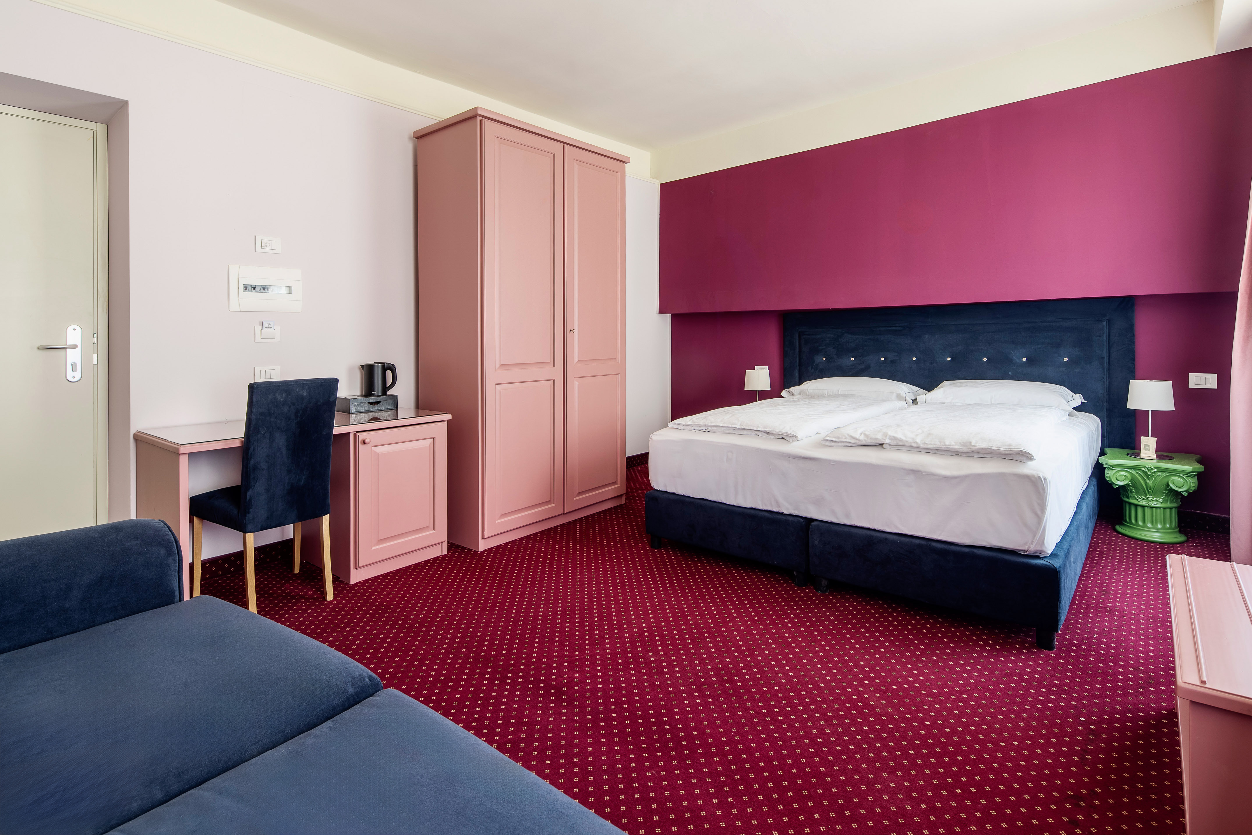 Magenta and pink hotel room with blue bed, red carpet, desk, and sofa.