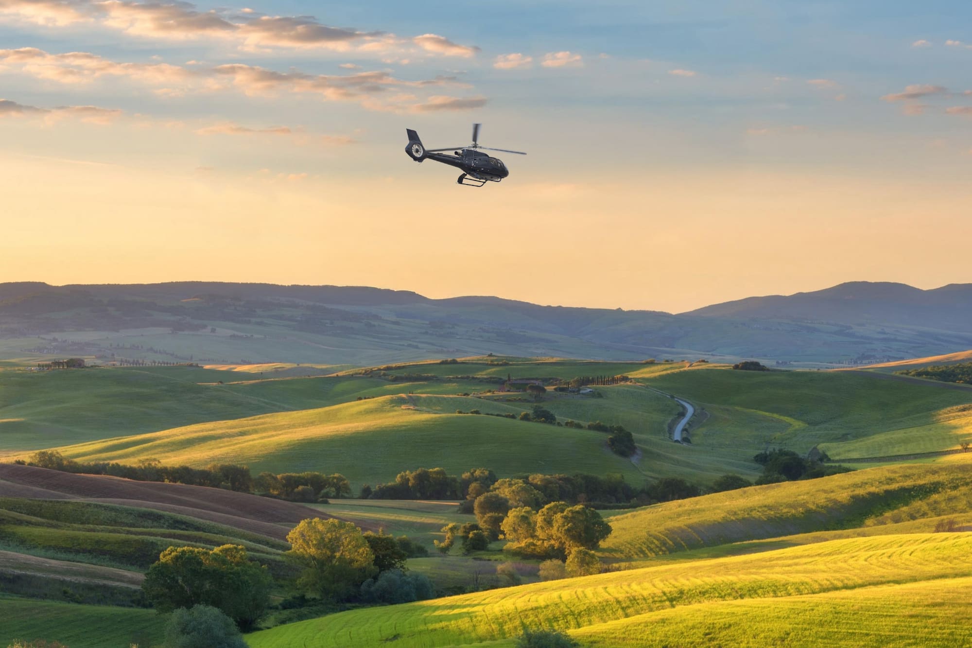 Helicopter flying above green rolling hills, golden sunset. Tuscany landscape.