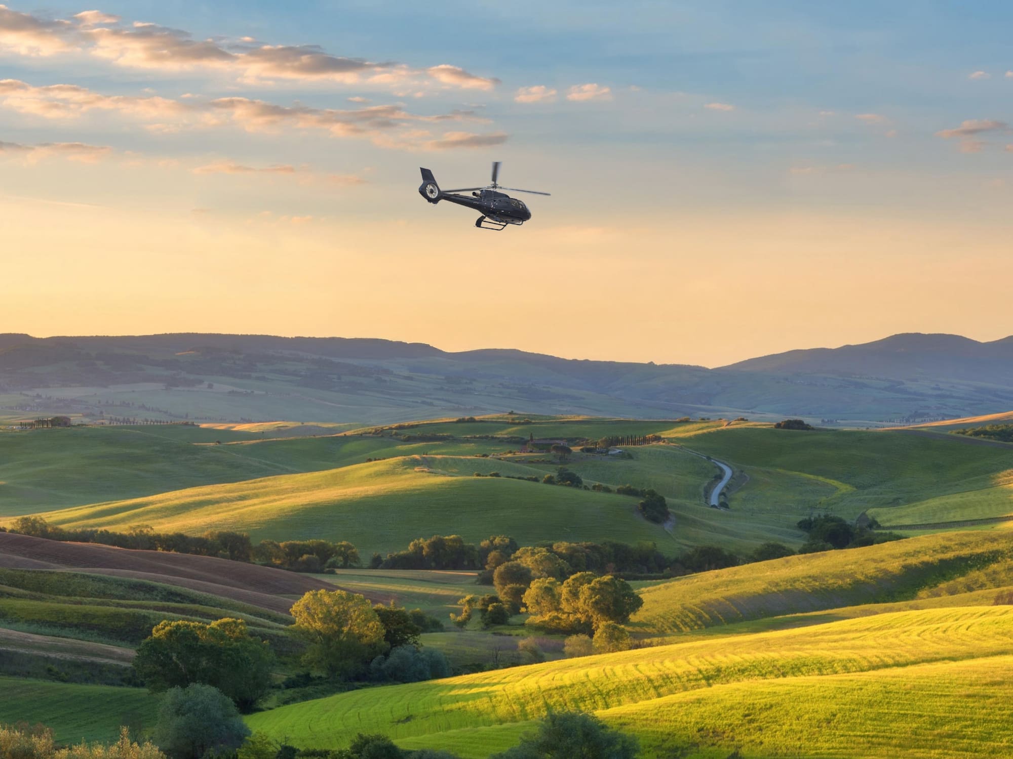 Helicopter flying above green rolling hills, golden sunset. Tuscany landscape.