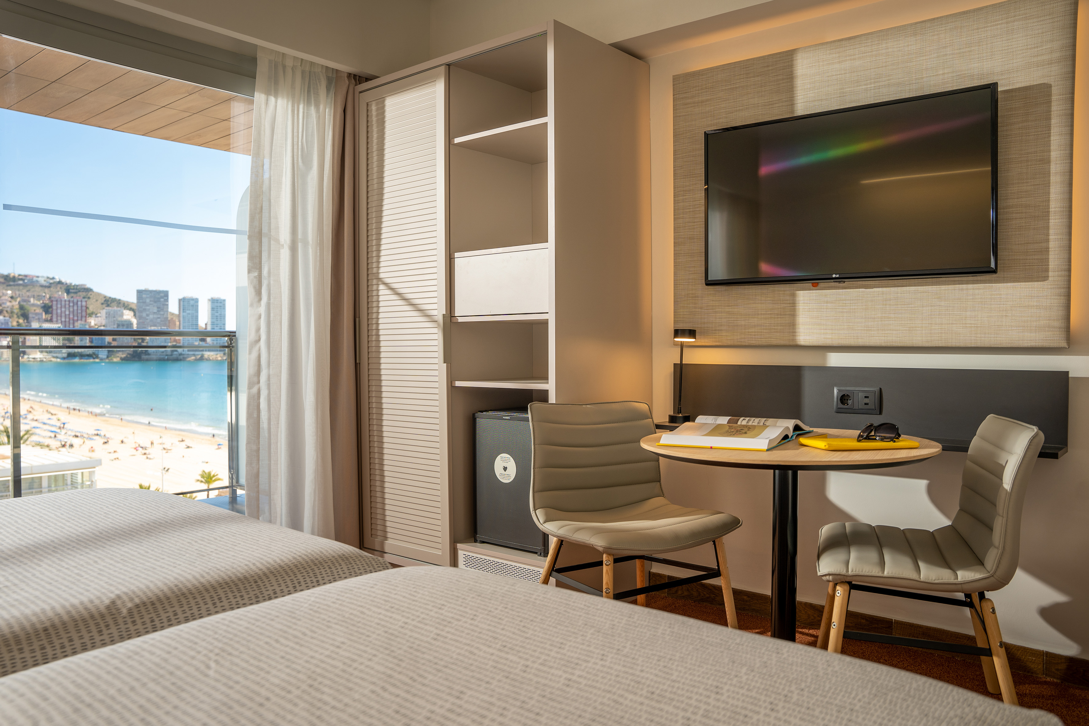 Bright modern hotel room with twin beds, ocean view balcony, and TV.