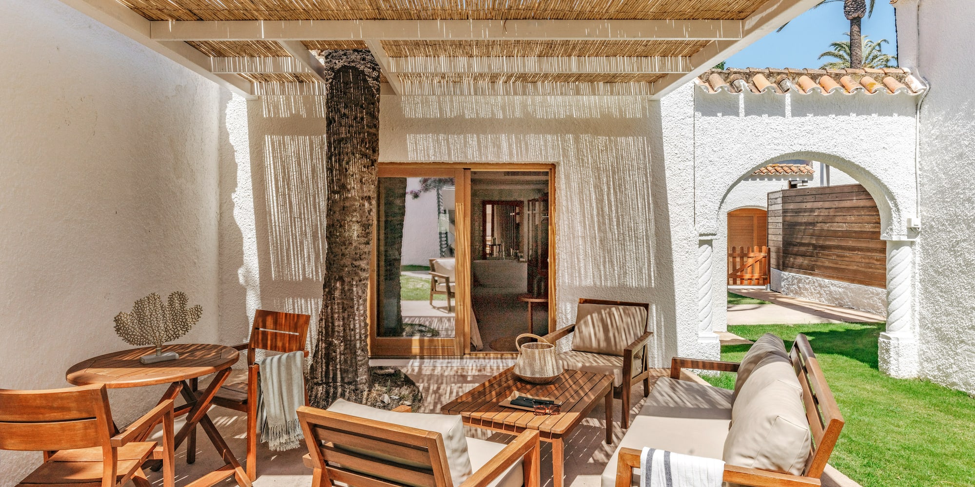 Sunny resort patio with wooden furniture, rattan canopy, and palm tree.