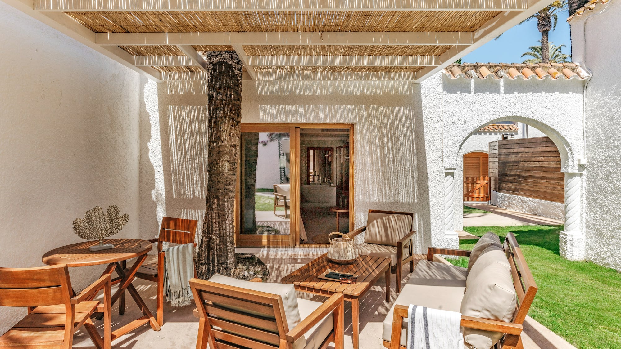 Sunny resort patio with wooden furniture, rattan canopy, and palm tree.