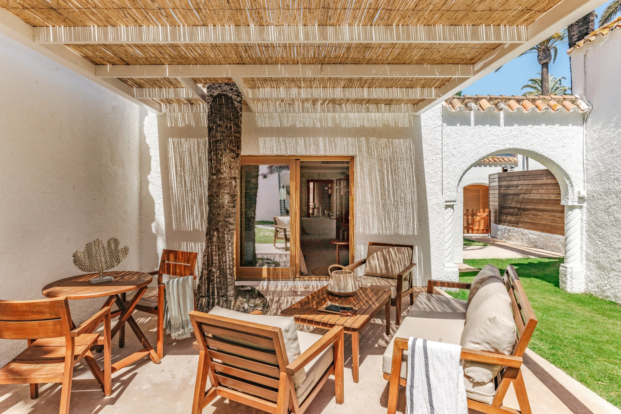 Sunny resort patio with wooden furniture, rattan canopy, and palm tree.