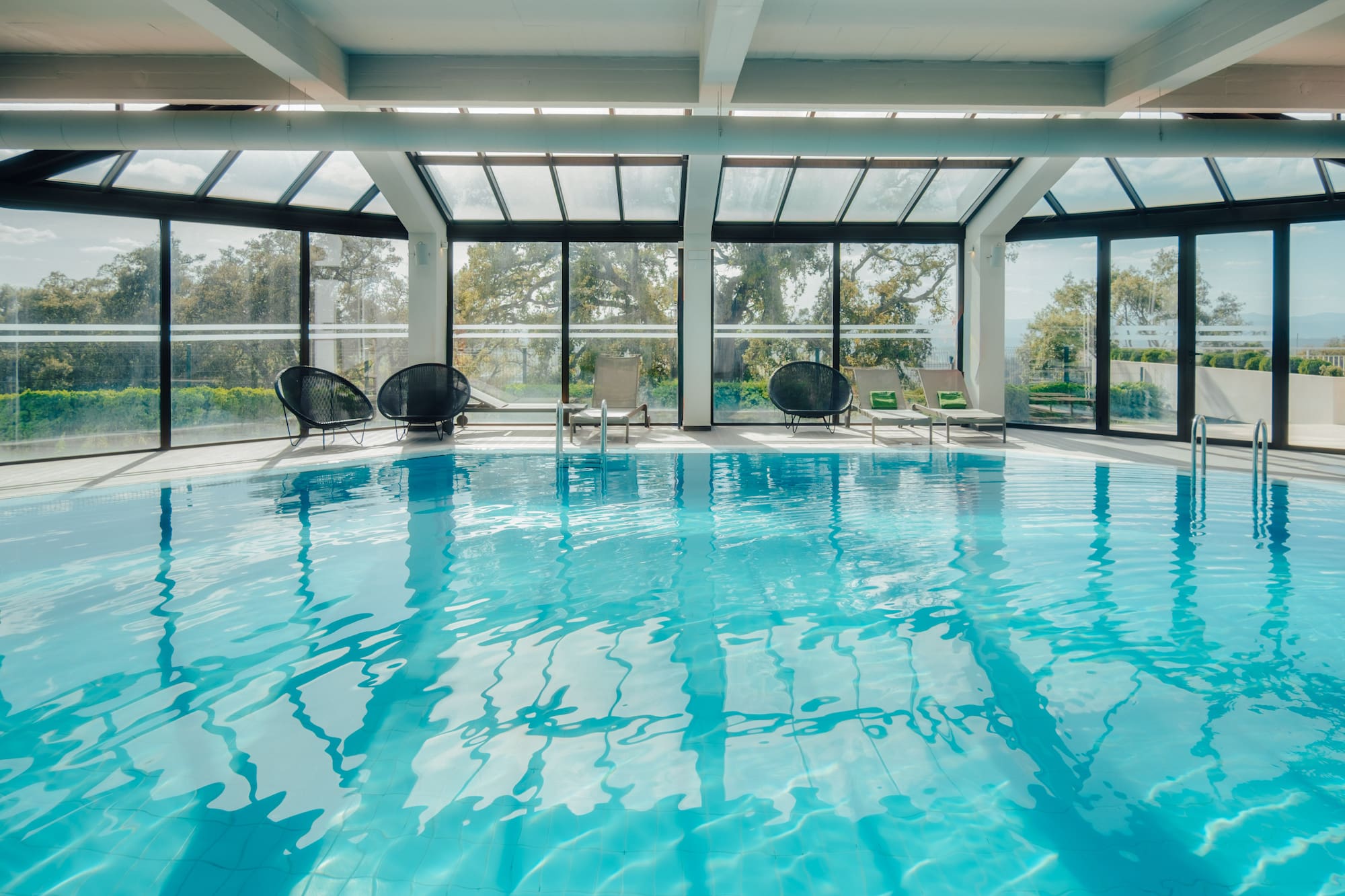 Indoor pool with glass roof, sun loungers, and expansive green outdoor view.