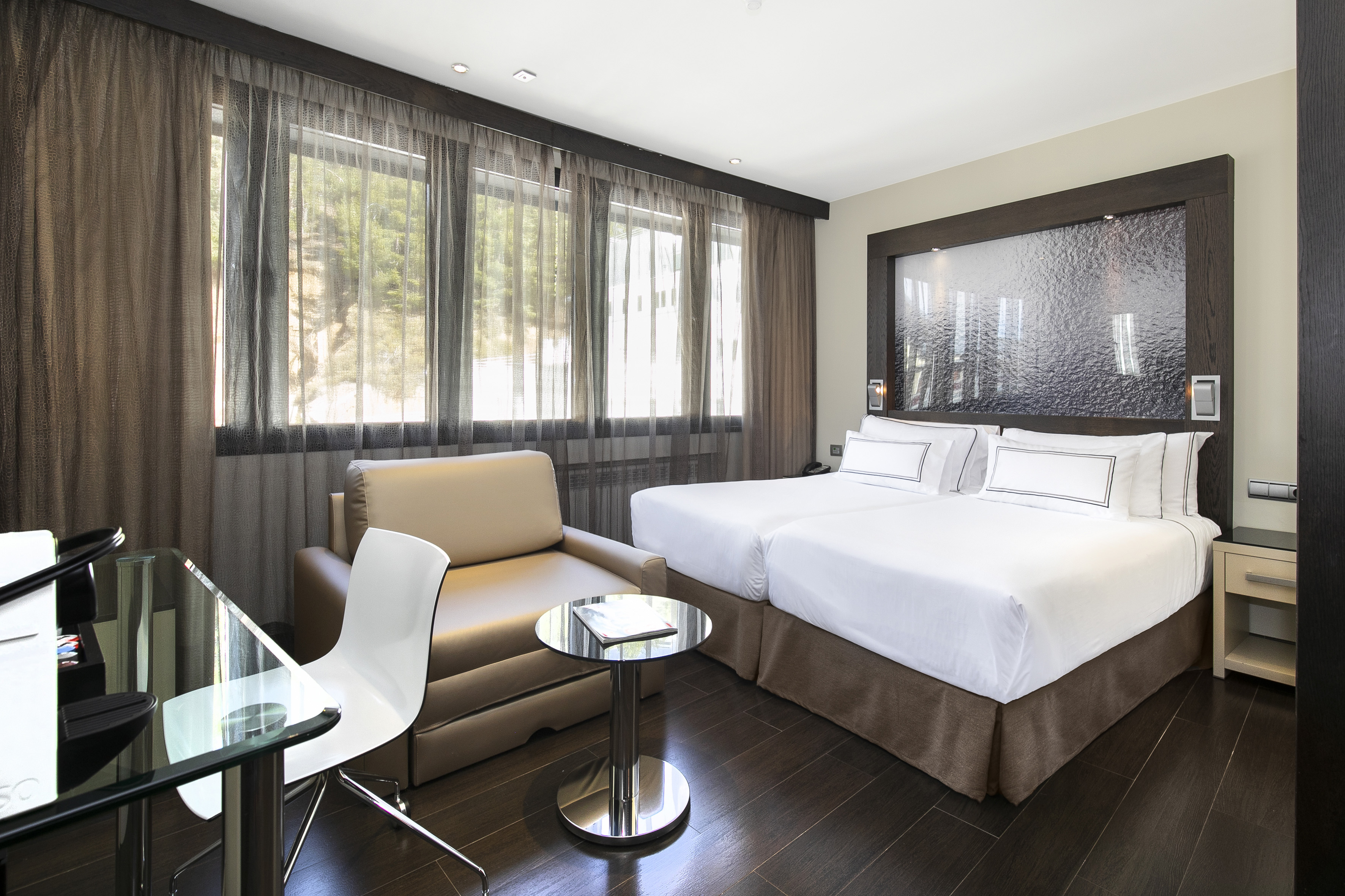 Modern hotel room with twin beds, sofa, glass desk, and sheer curtains.