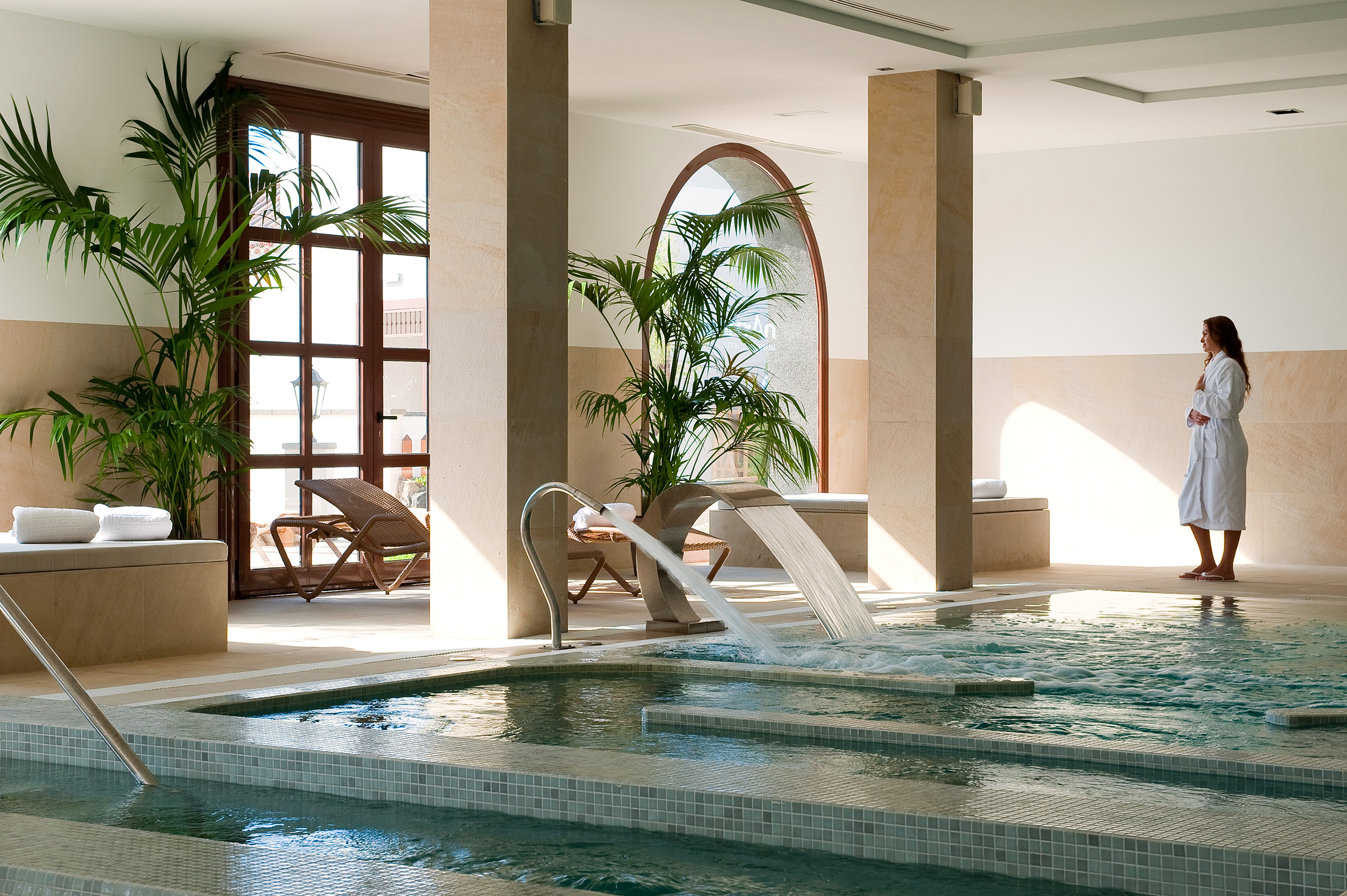 Tropical indoor spa pool with waterfall, palm trees, and woman in robe.