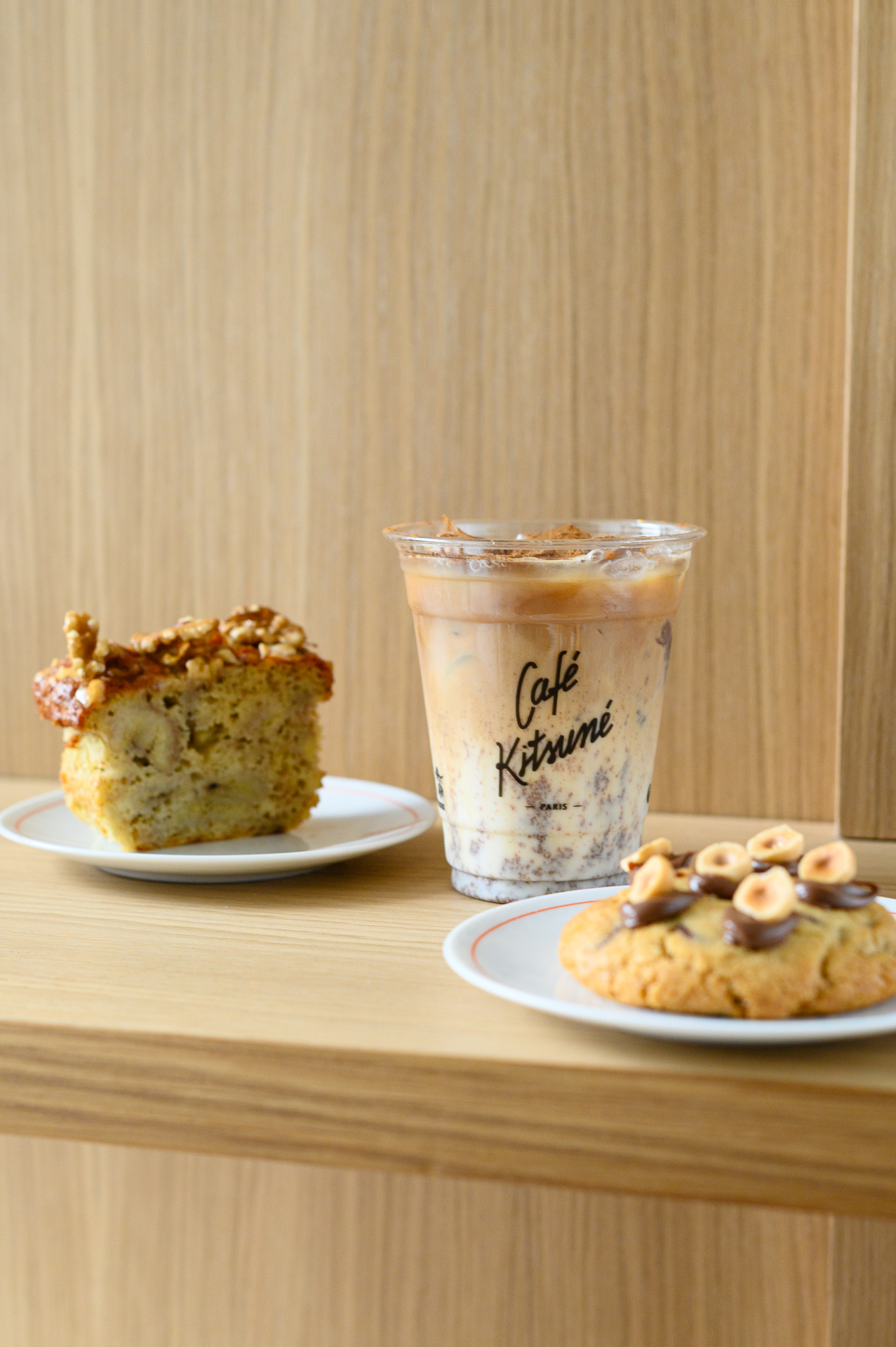 Café Kitsuné iced latte, banana bread with walnuts, and hazelnut cookie on wood.