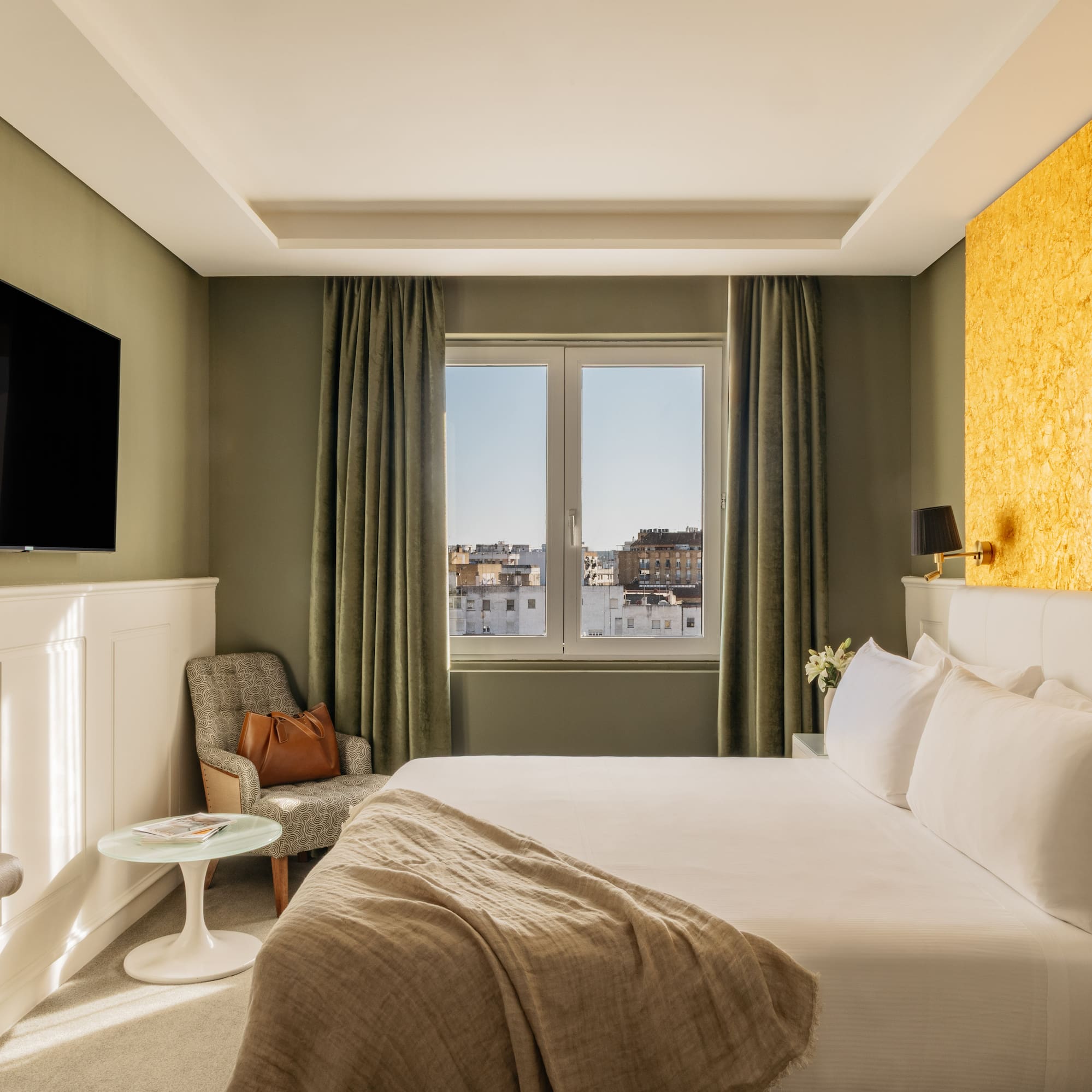 Moderna habitación de hotel con cama, TV, pared dorada y vista a la ciudad.