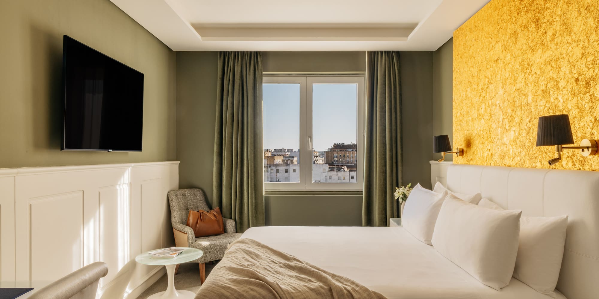 Moderna habitación de hotel con cama, TV, pared dorada y vista a la ciudad.