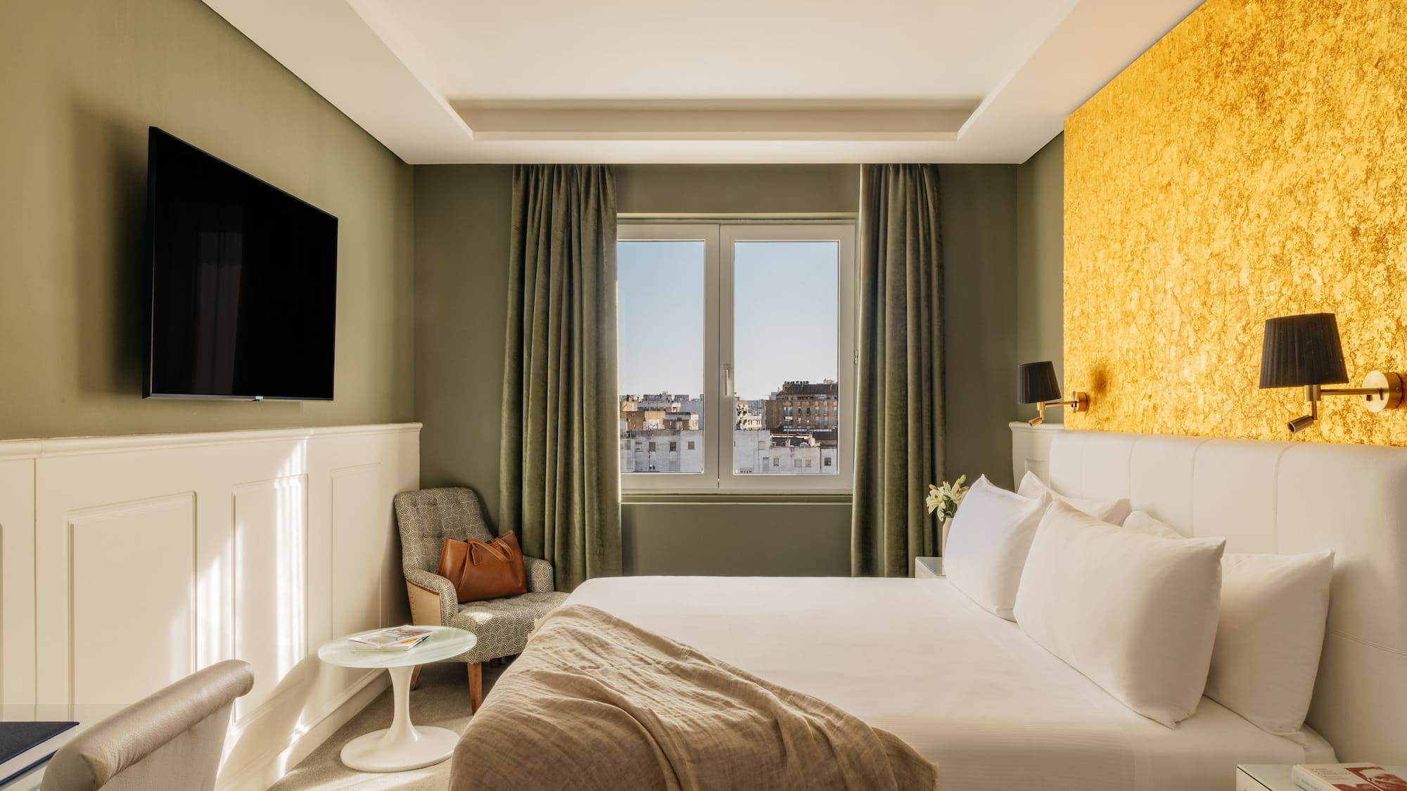 Moderna habitación de hotel con cama, TV, pared dorada y vista a la ciudad.