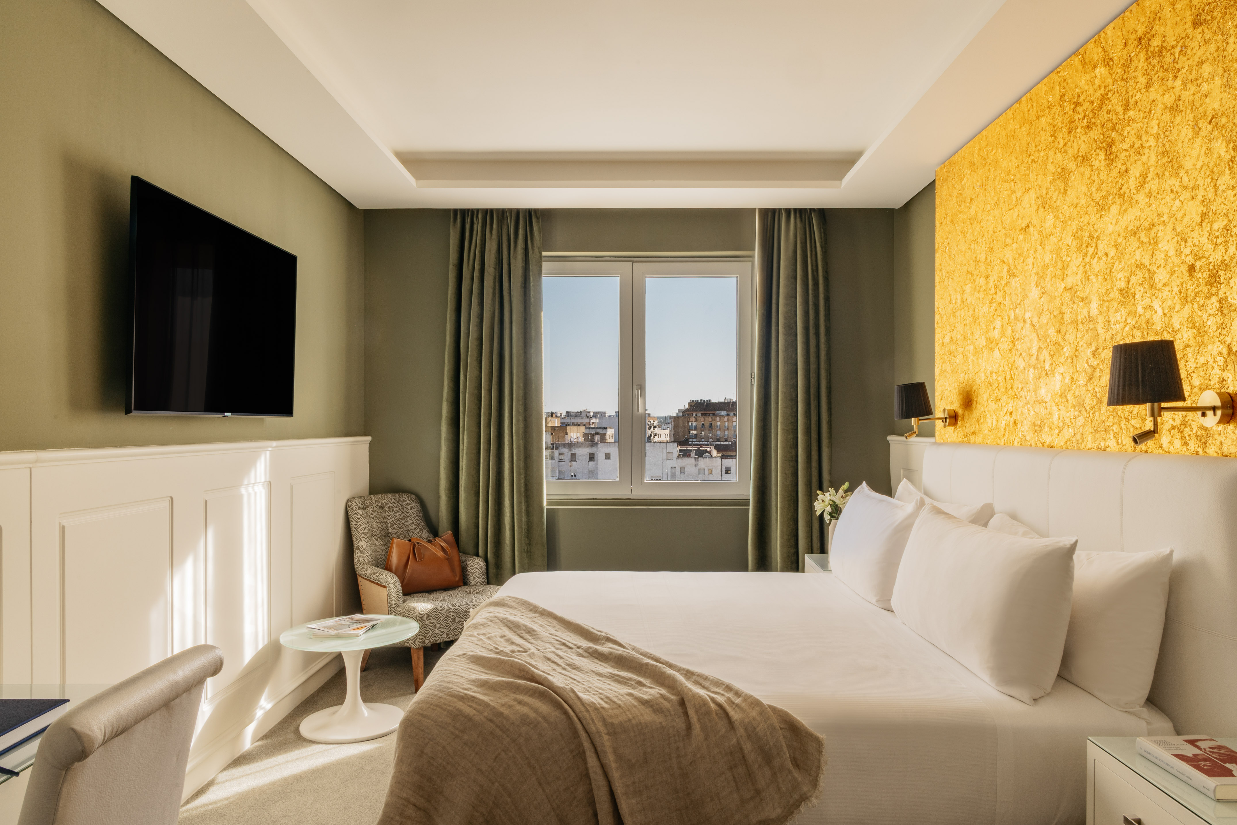 Moderna habitación de hotel con cama, TV, pared dorada y vista a la ciudad.