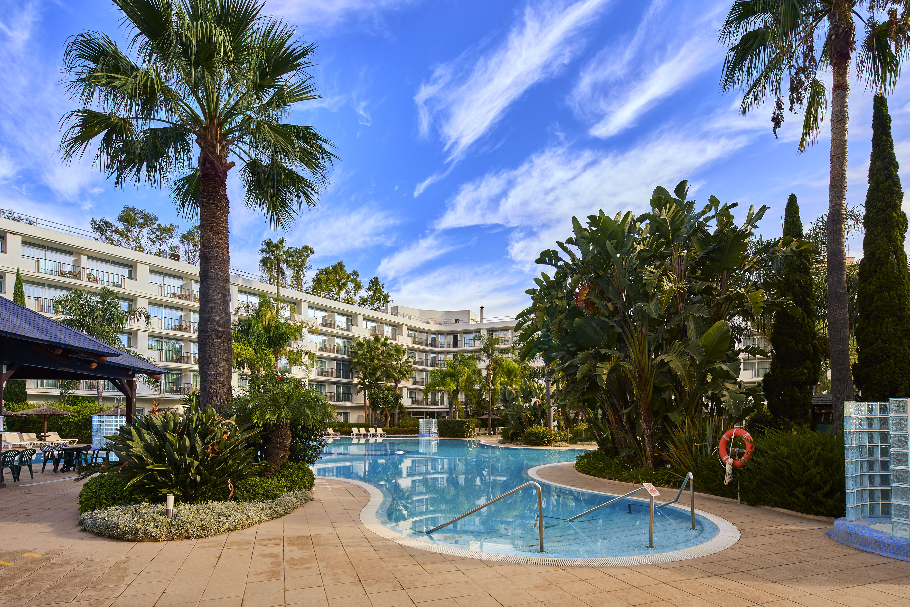 Sunny resort swimming pool with palm trees and hotel under a blue sky.